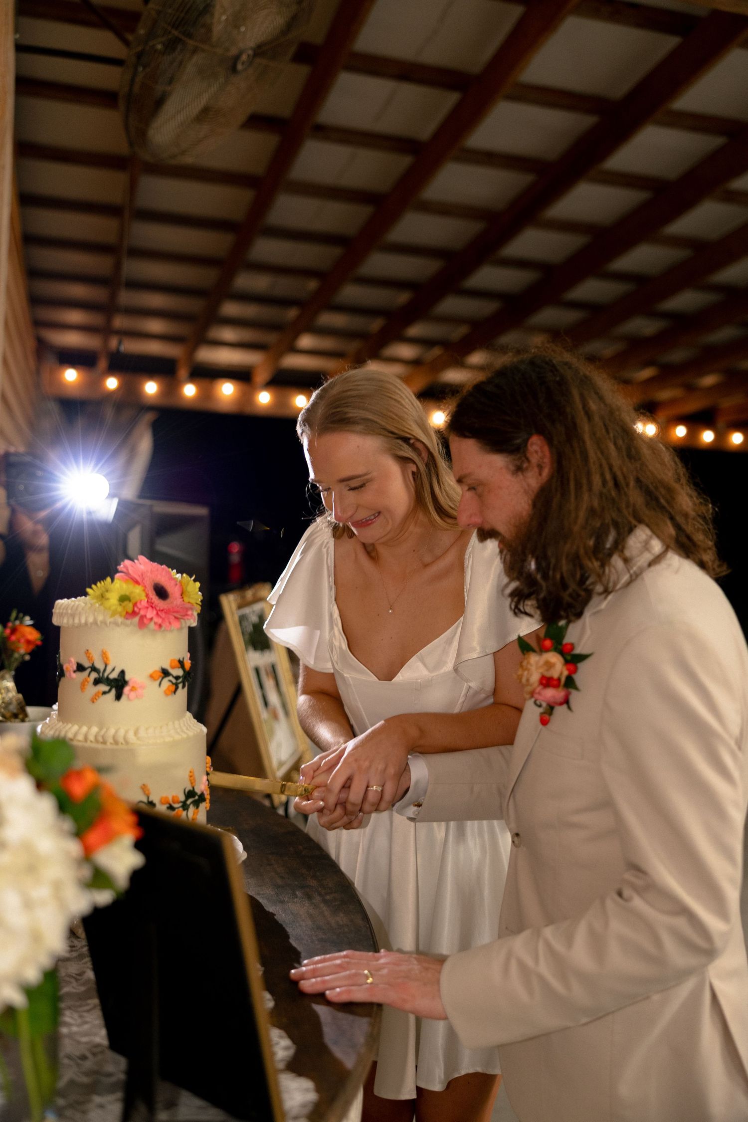 Smiling bride and groom in white attire cut floral wedding cake at rustic barn reception.