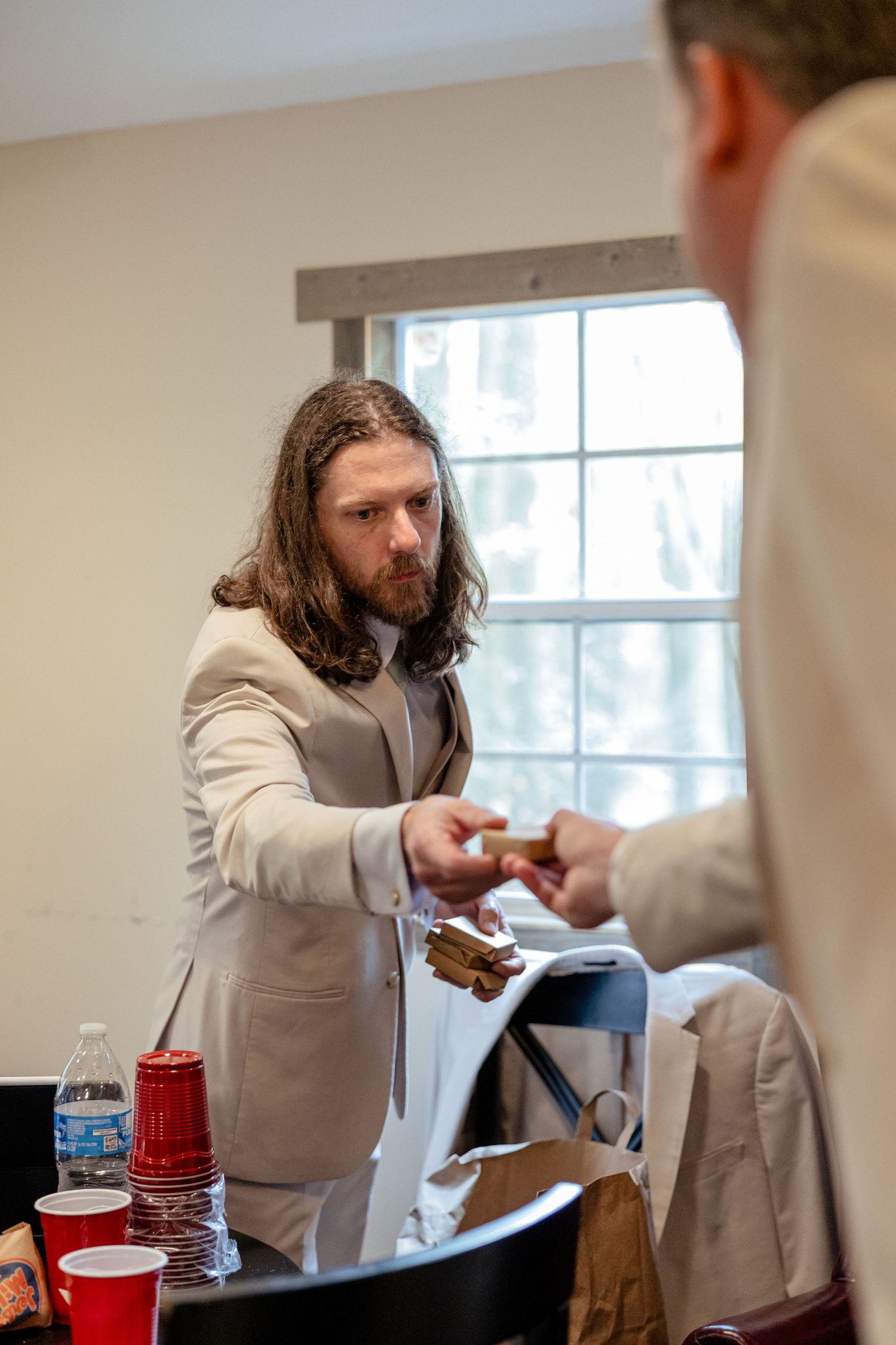 Long-haired groomsman in beige suit exchanges gift with another man at a wedding preparation table.