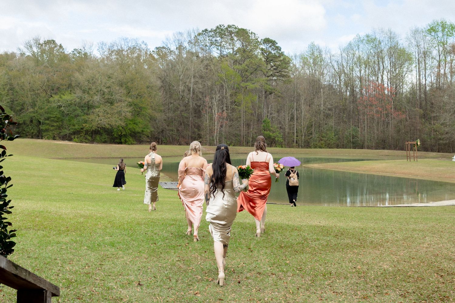 Bridesmaids in colorful dresses running across green lawn toward a pond at an outdoor wedding venue.
