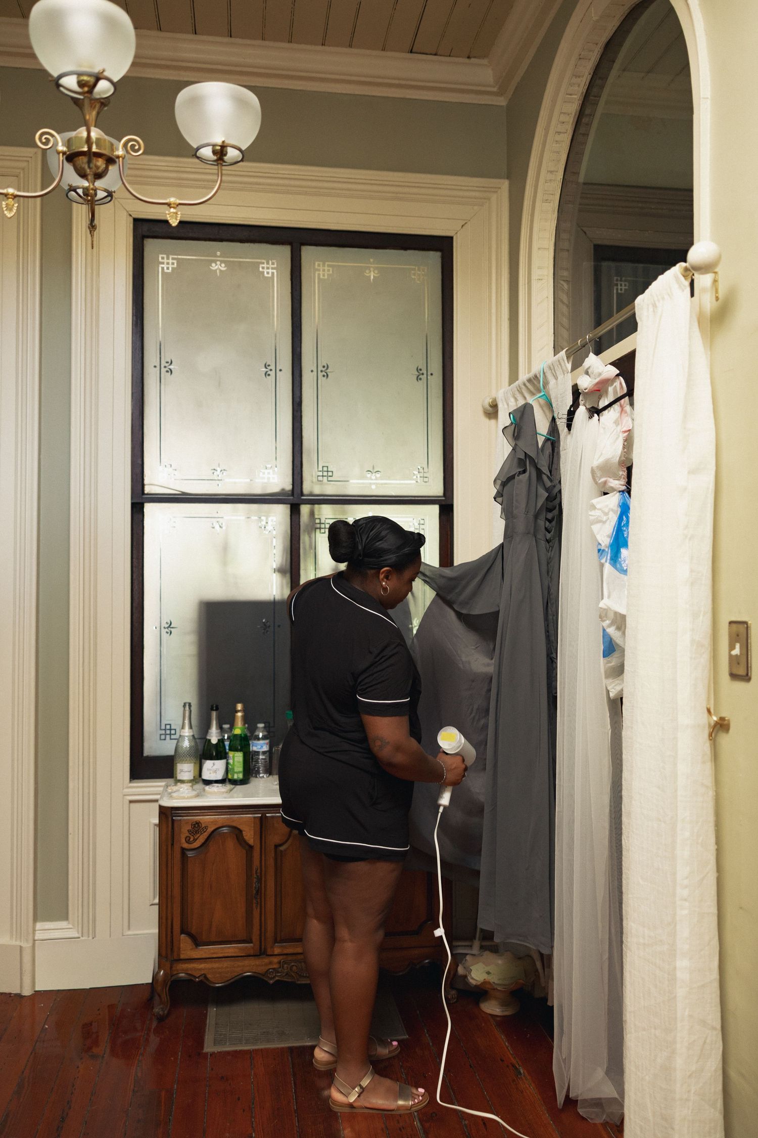A hotel maid in uniform steams a gray dress hanging near a window in an elegant room with hardwood floors.