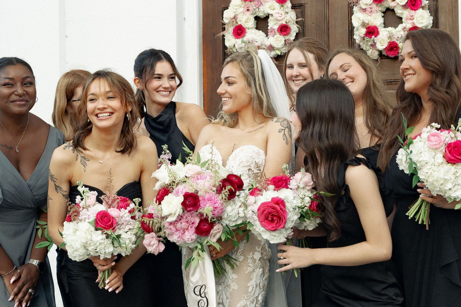 Smiling bride in lace gown and veil surrounded by bridesmaids in black dresses with colorful bouquets.