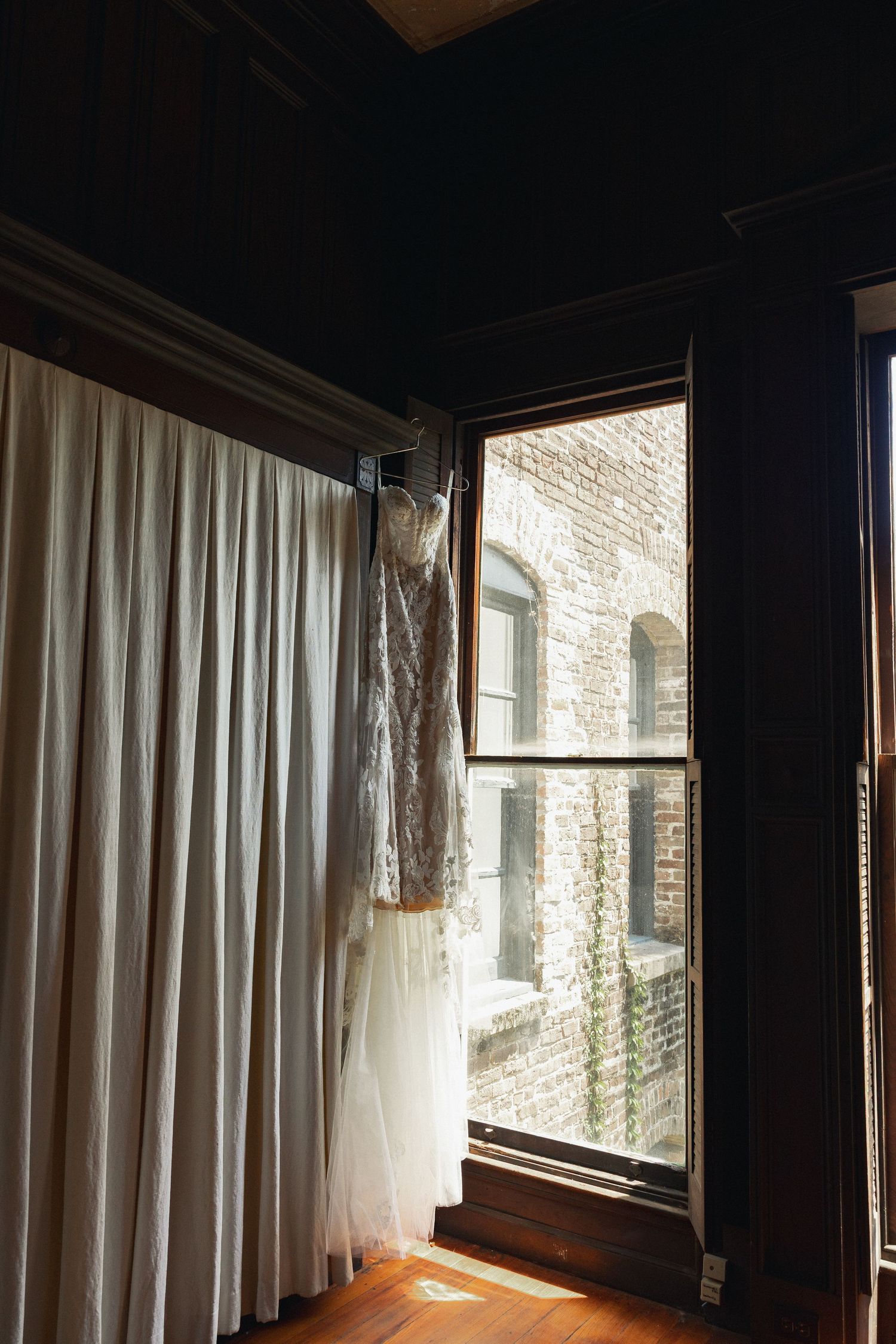 Elegant wedding gown hanging near a window with brick wall view.