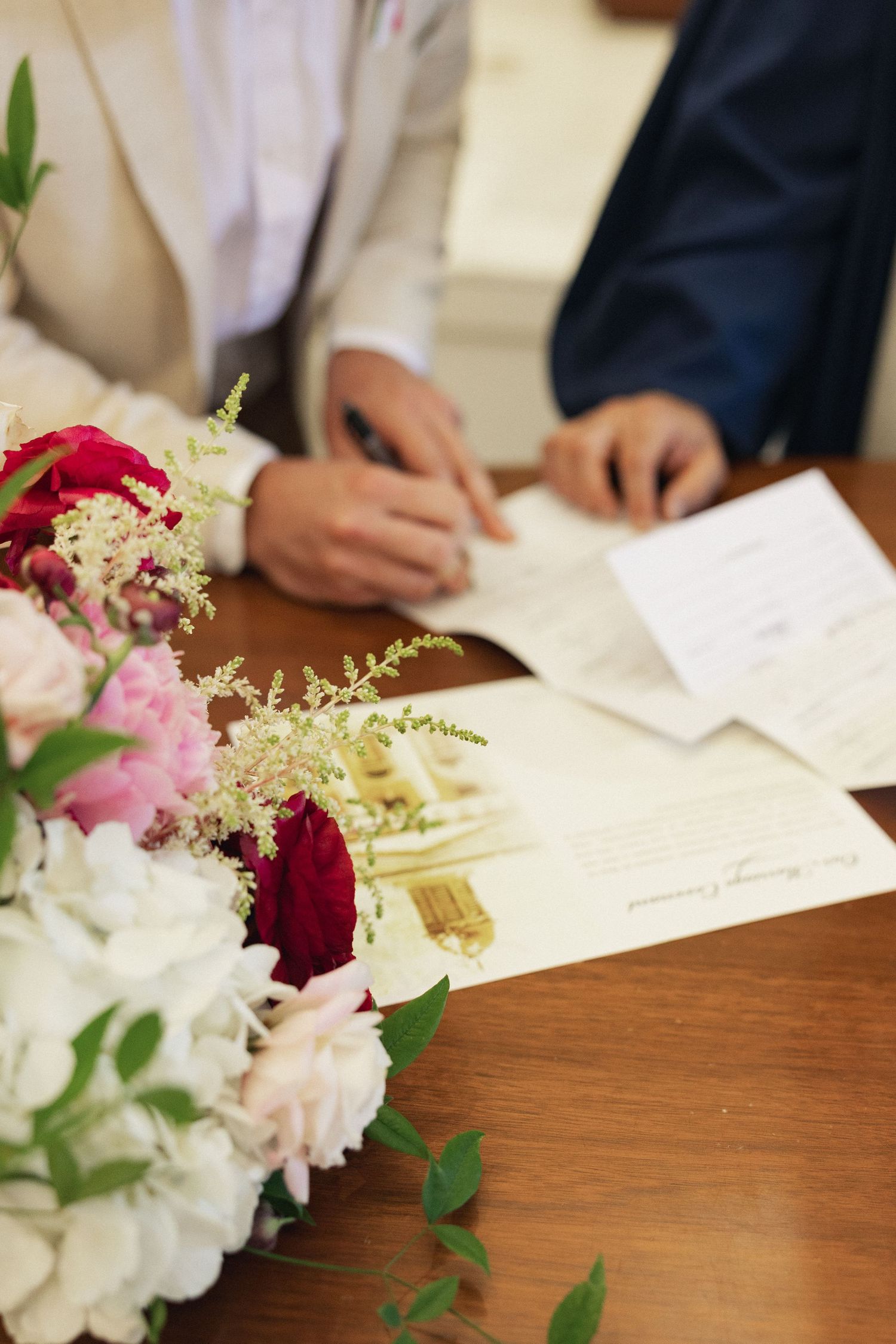Couple signing wedding certificate at wooden table with bridal bouquet of red and white flowers in foreground.