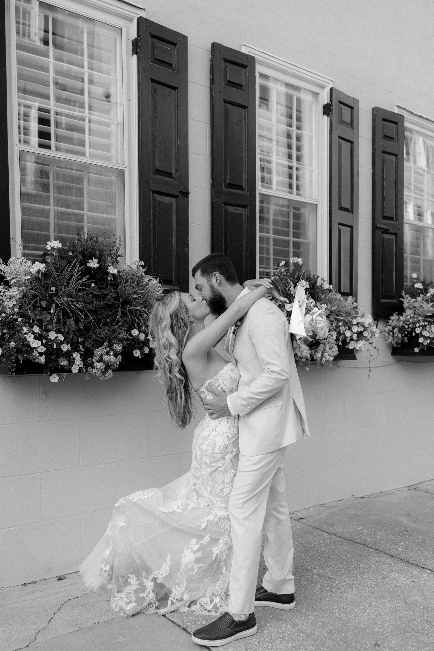 Black and white photo of bride and groom sharing romantic kiss before window boxes filled with flowers on city street.