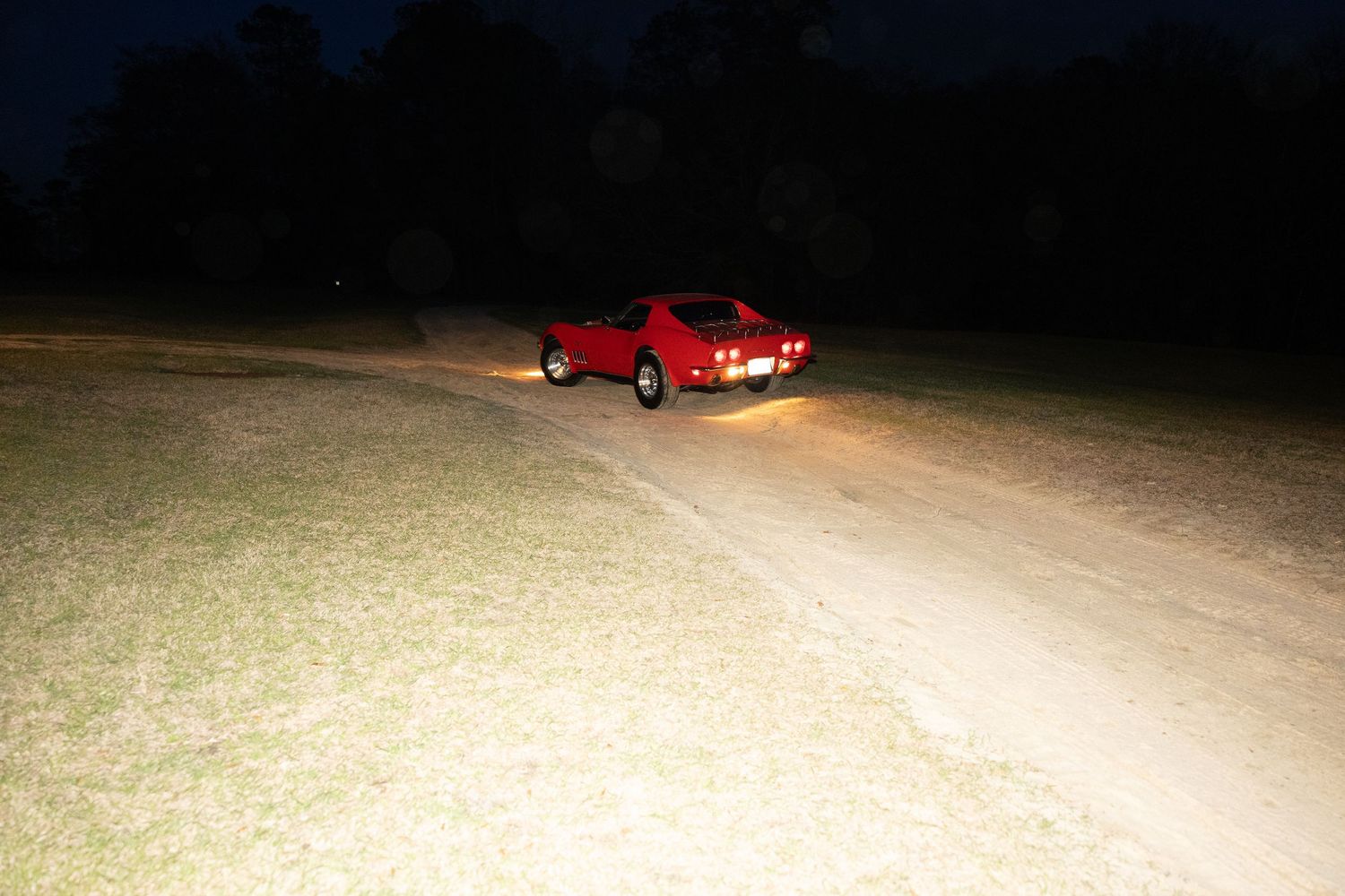 Red sports car driving on a dirt path at night with headlights illuminating the ground in a dark open field.