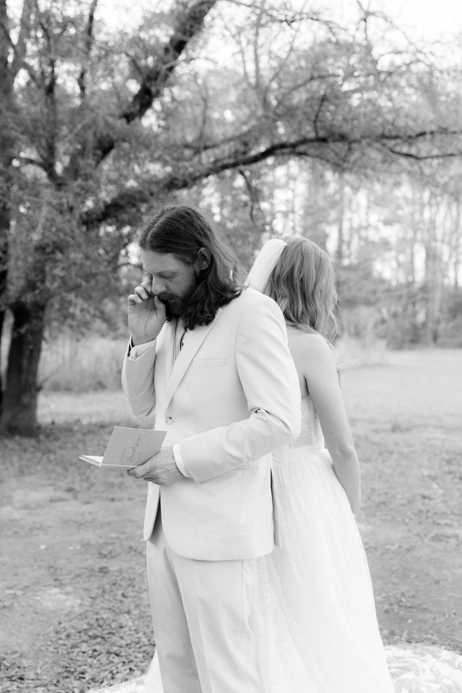 Emotional groom in white suit reads vows while standing back to back with bride under bare tree in black and white.