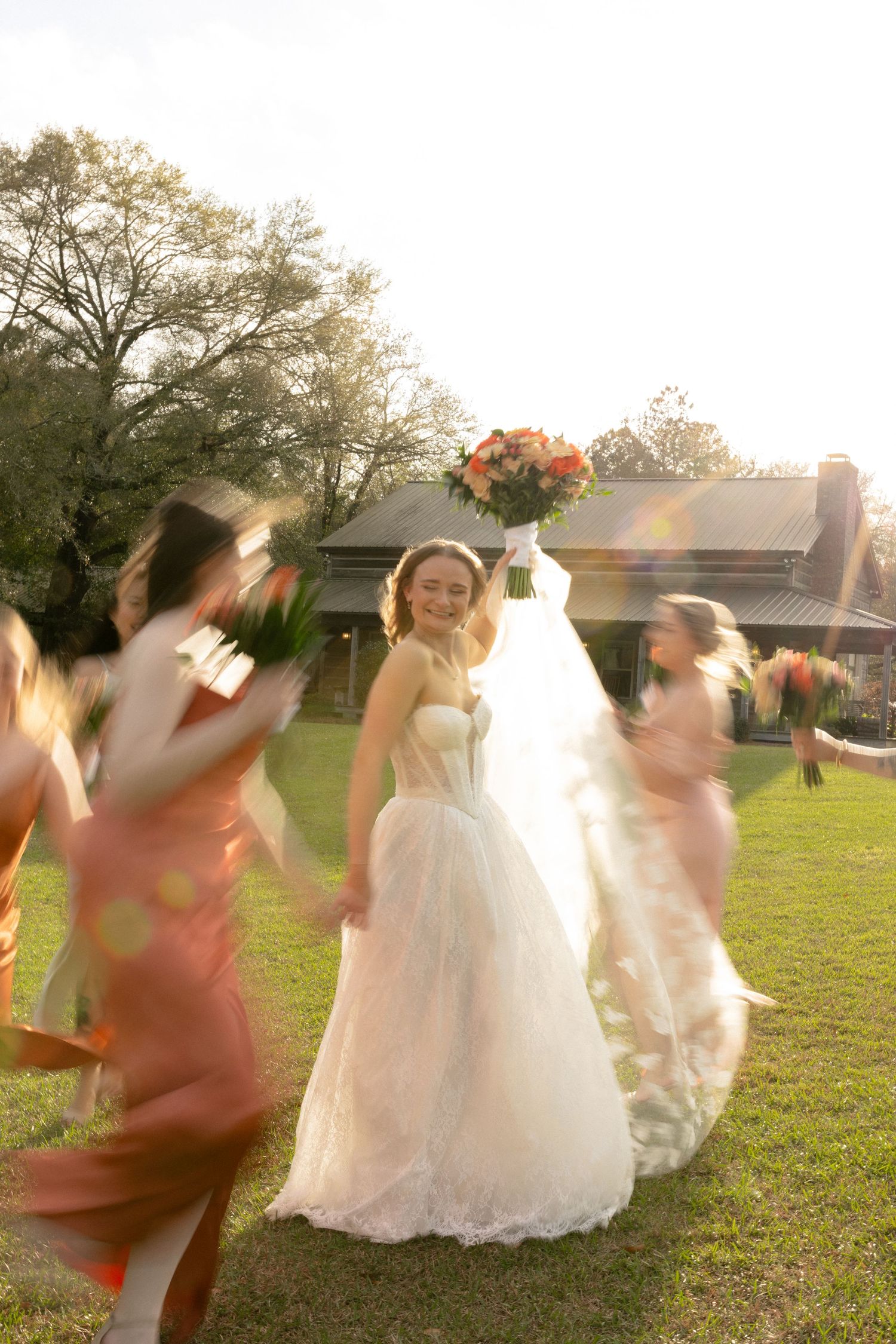 Bride raising orange bouquet joyfully while bridesmaids in coral dresses swirl around her on green lawn.