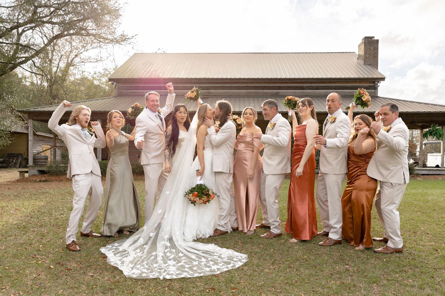 Joyful wedding party celebrates with raised arms outside sunlit rustic barn venue.