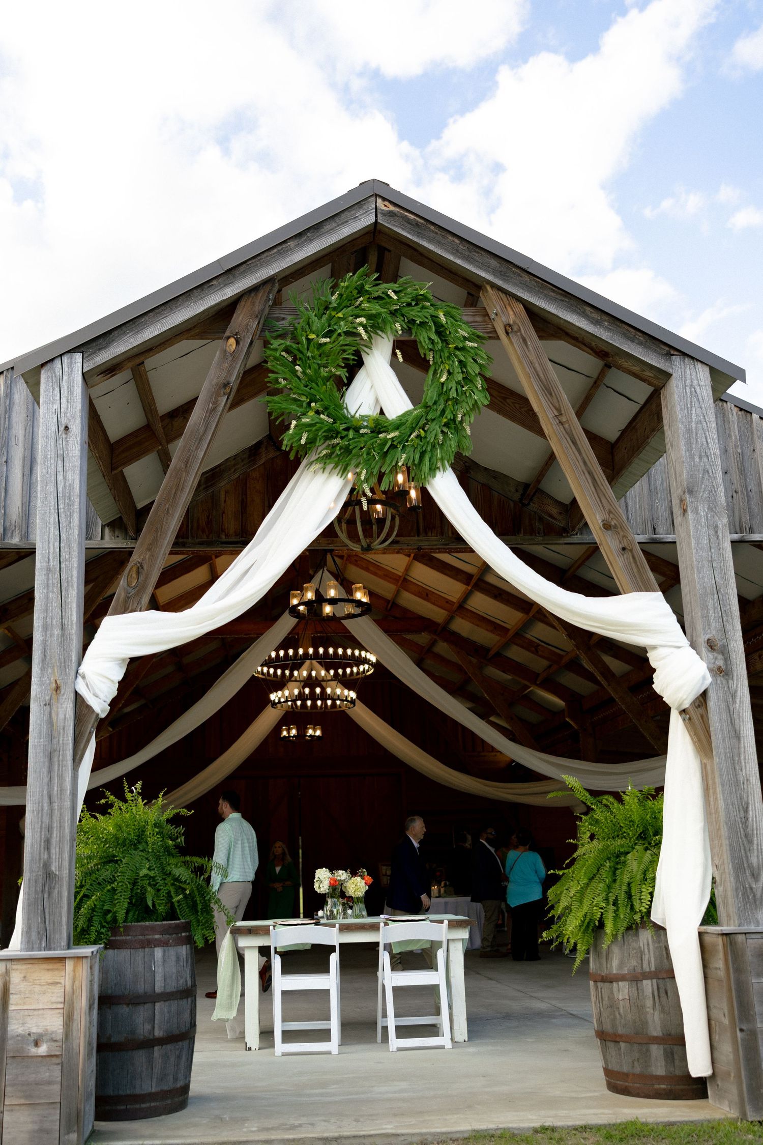 Rustic barn wedding venue entrance decorated with white draping, a green wreath, and barrel planters.