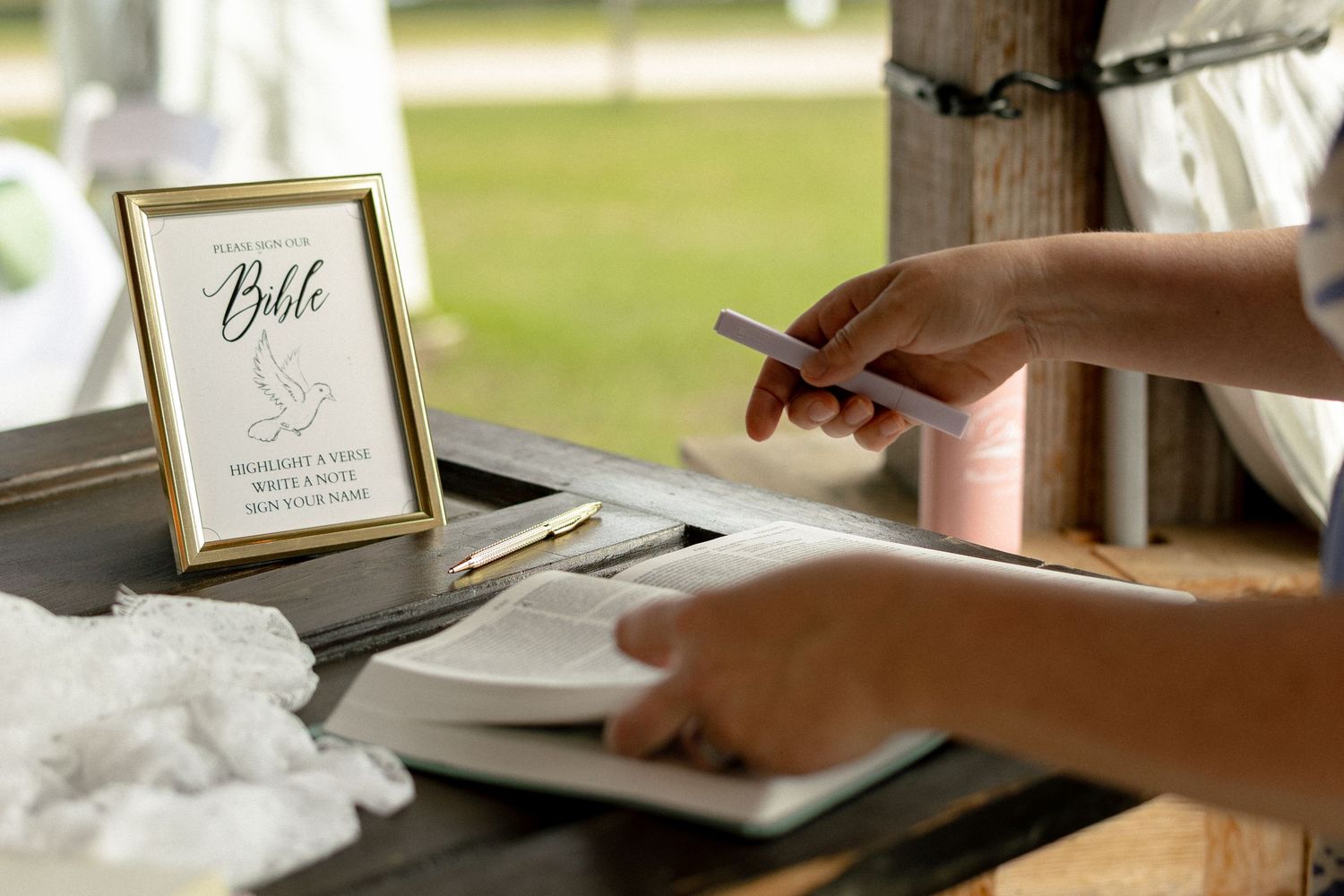 Guest signing a Bible at a rustic wedding reception table with gold-framed sign and white lace decor.