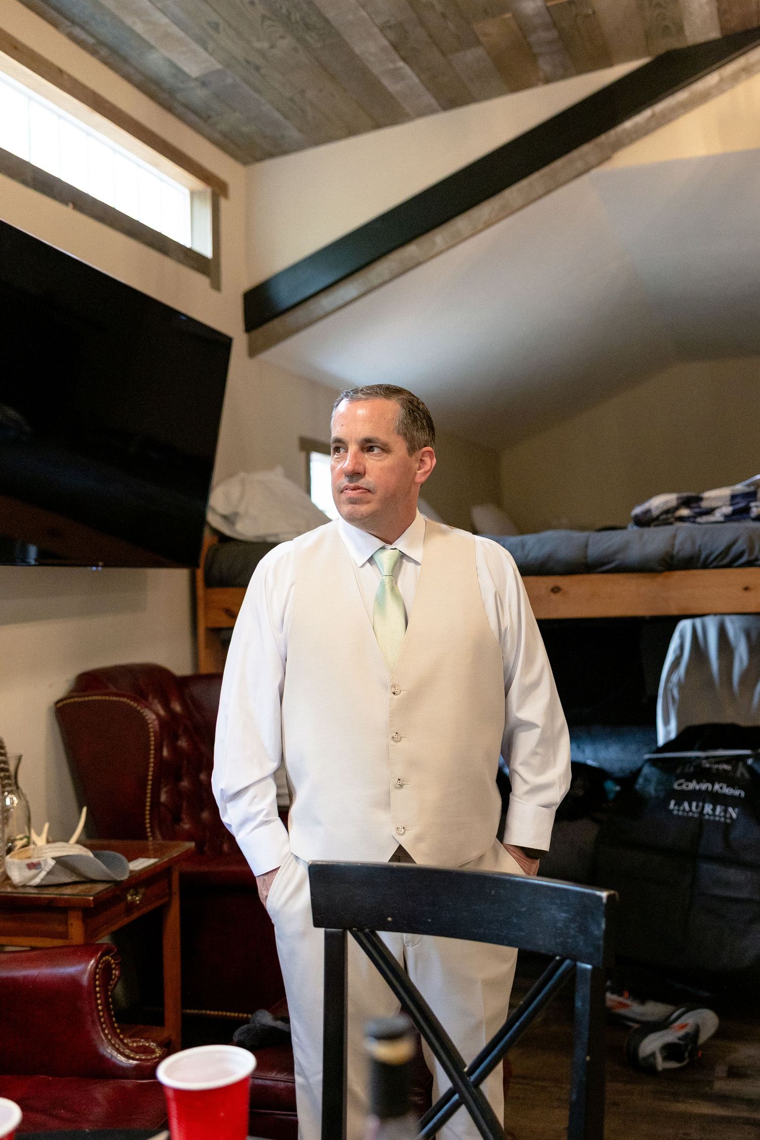 Groom in white vest and mint tie standing in rustic wood-ceiling room during wedding preparations.