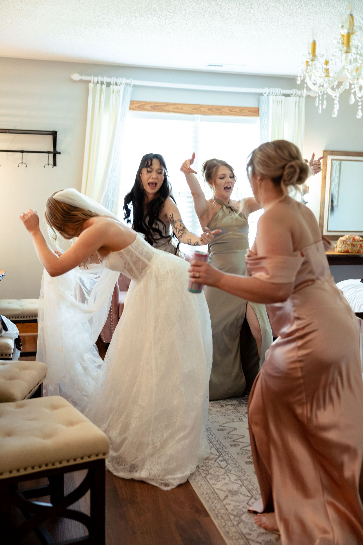 Excited bridesmaids cheering around bride in lace wedding gown during fun getting-ready moment.