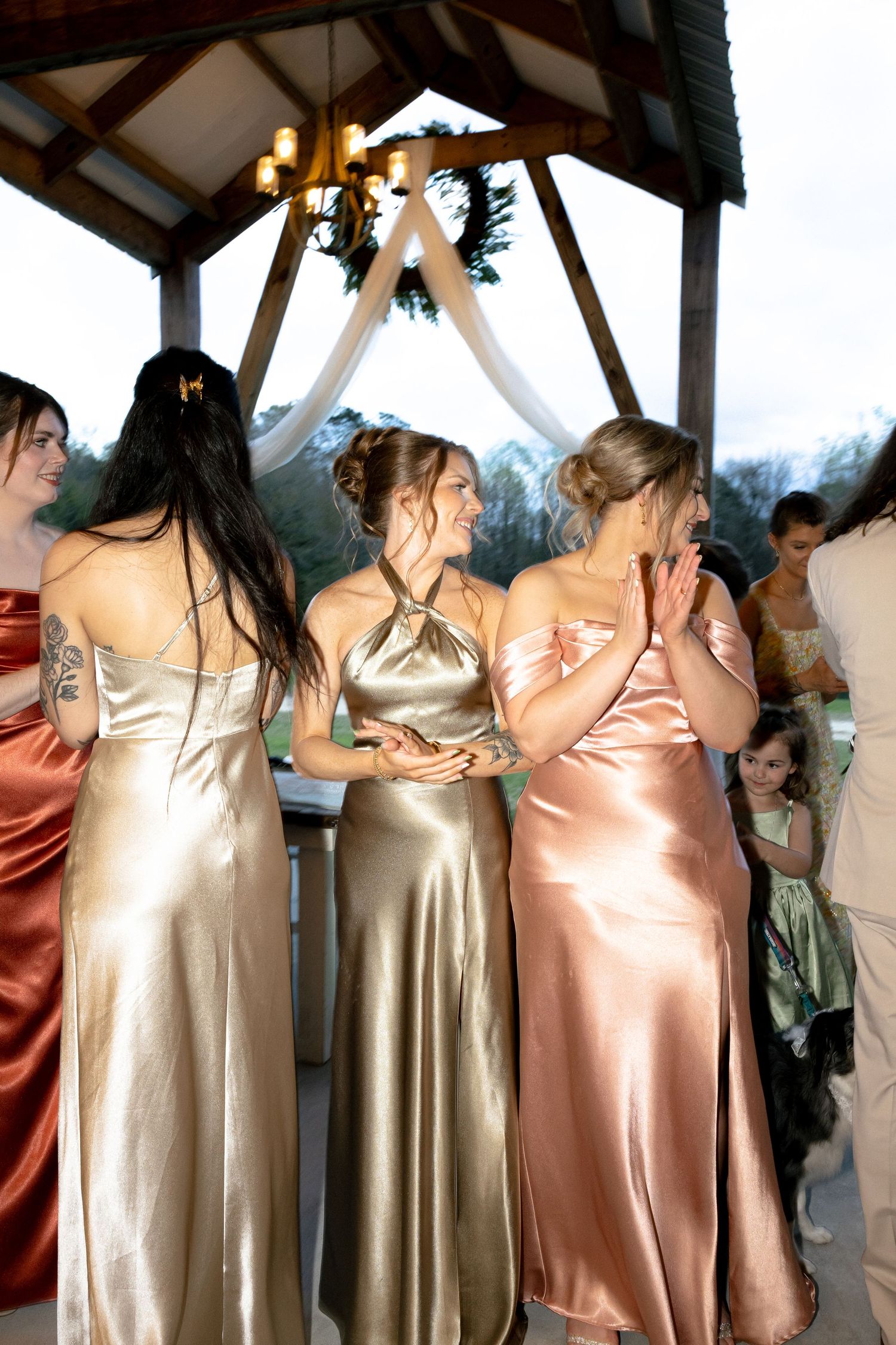 Bridesmaids in satin gowns of gold and rust stand together laughing under a gazebo at outdoor wedding.