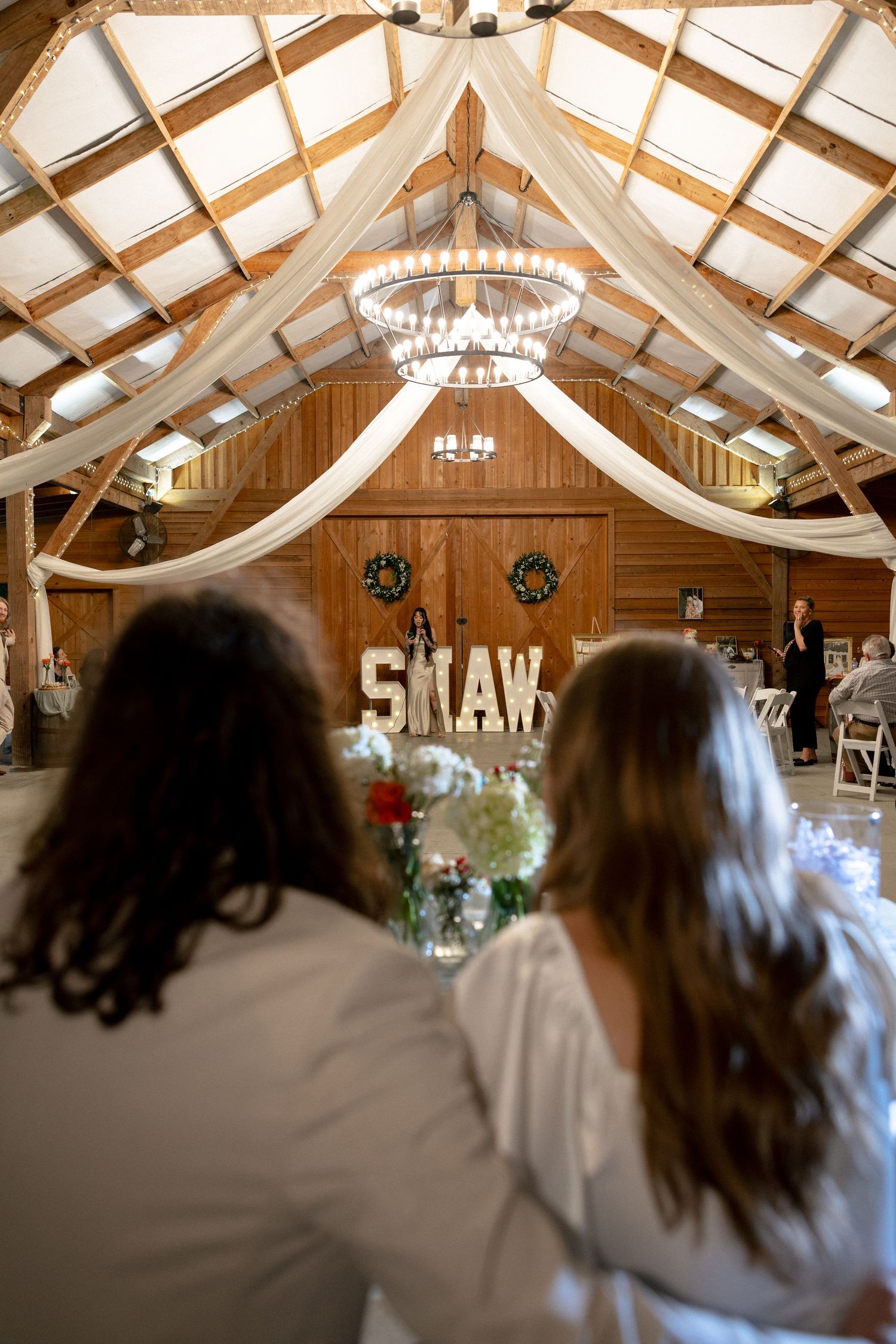Rustic barn wedding venue with white draping, chandelier, and illuminated SHAW marquee letters.