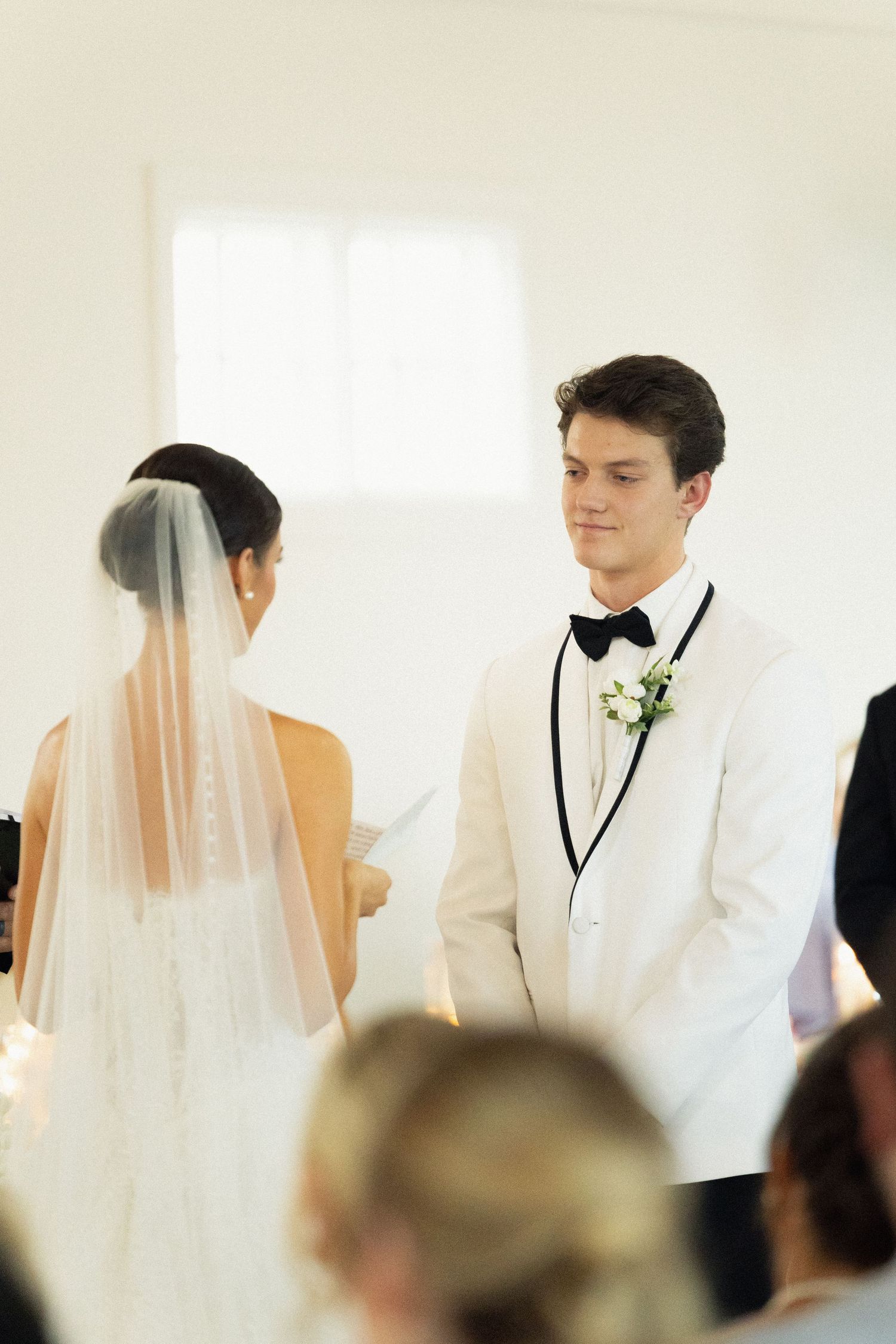 Groom in white tuxedo smiling at bride reading vows during wedding ceremony.