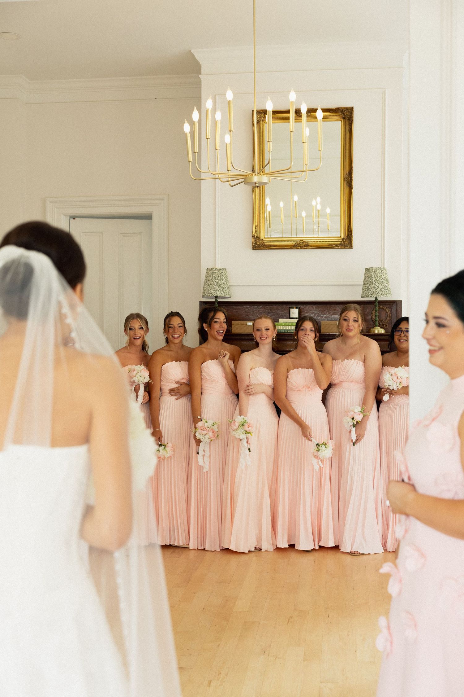 Bridesmaids in blush pink strapless gowns holding white bouquets react to bride in elegant white wedding venue.