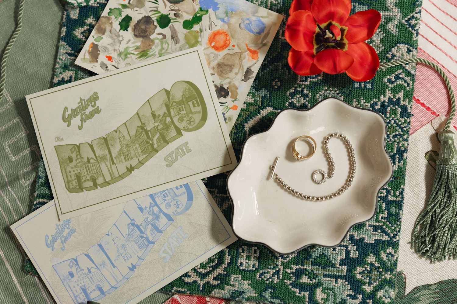 Flat lay of vintage postcards, pearl jewelry in a white dish, and a red flower on a green patterned cloth.