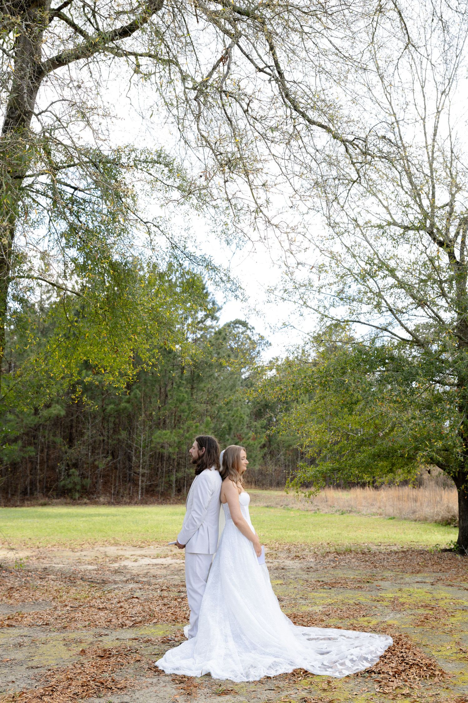 Bride and groom stand back-to-back under a large oak tree in an outdoor rustic wedding setting.