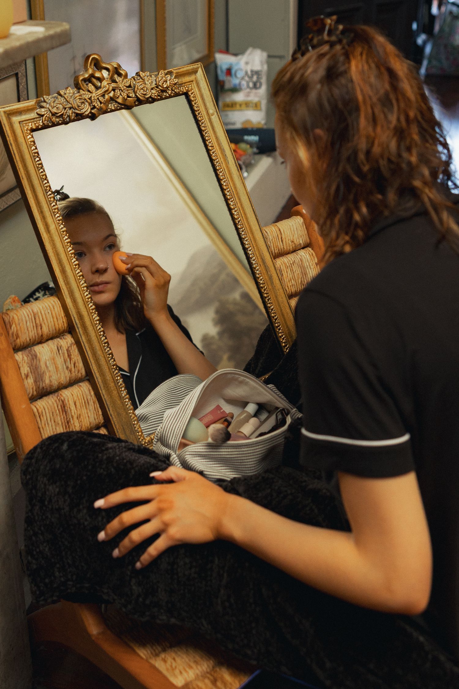Young woman applying makeup with a sponge while looking into an ornate gold-framed mirror, with a makeup bag nearby.
