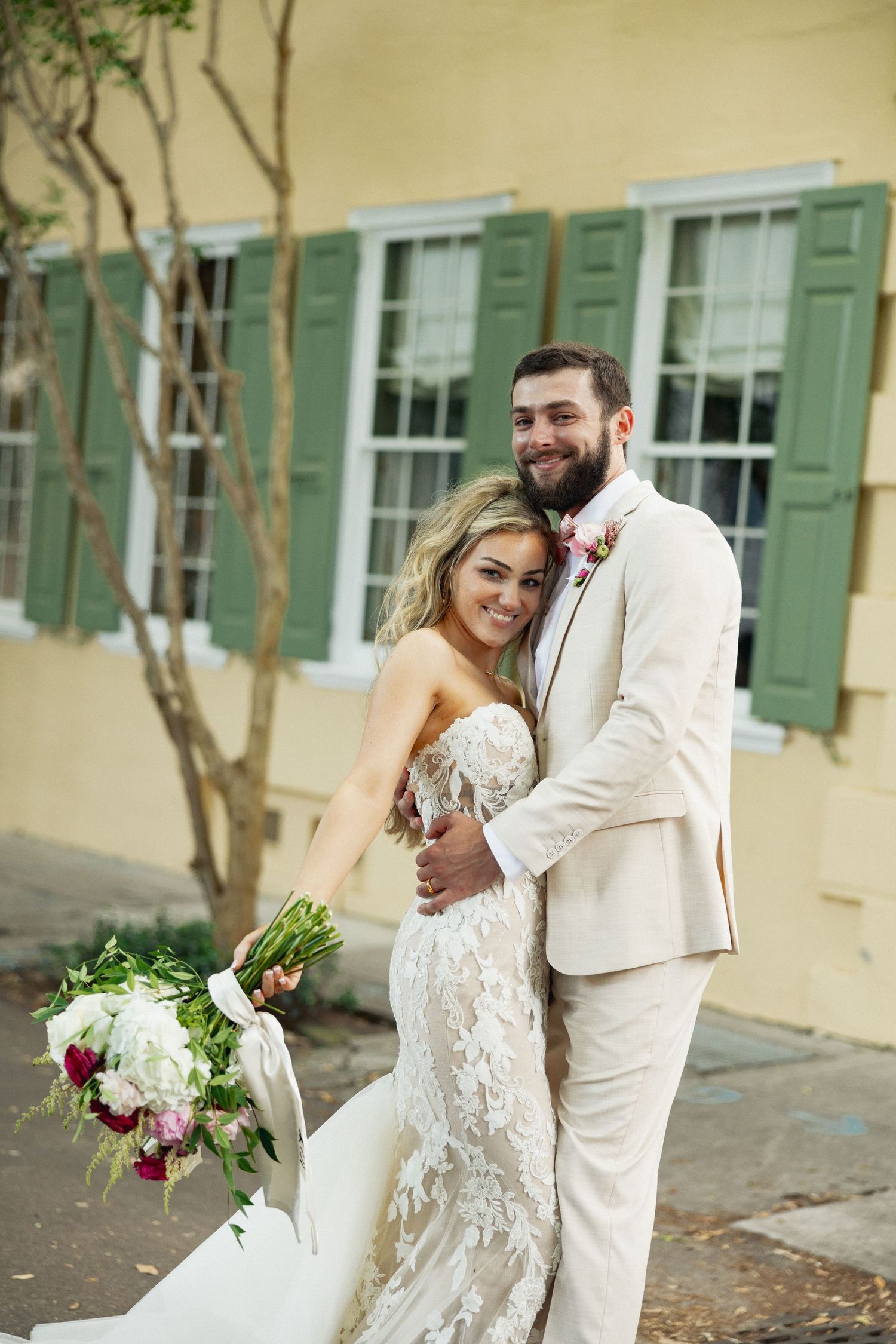 Smiling bride in lace mermaid gown and groom in cream suit embrace outside yellow building with green shutters.