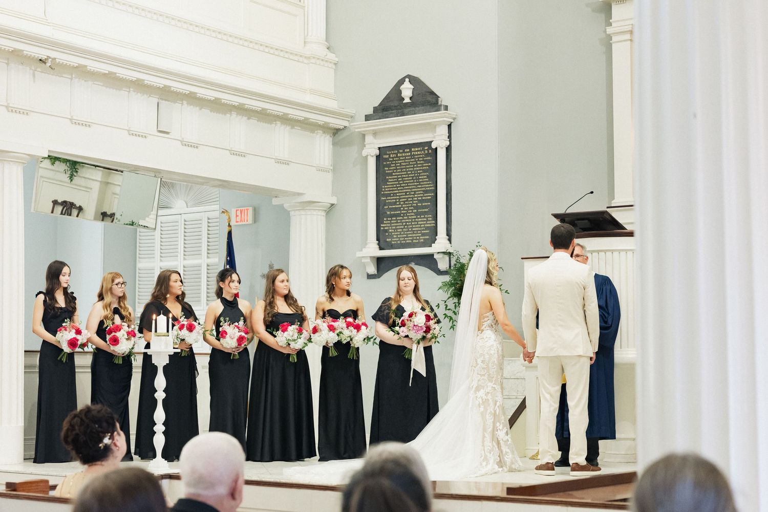 Bridesmaids in black gowns holding pink bouquets stand at altar during church wedding ceremony.