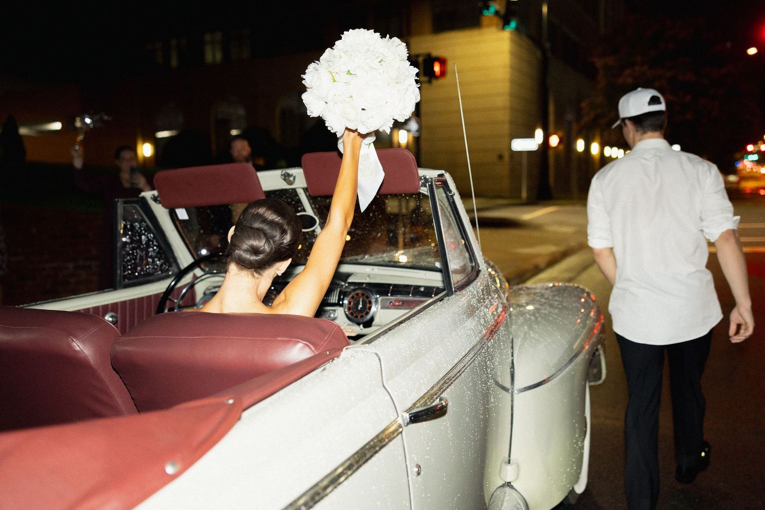 Bride in convertible classic car raises white bouquet at night, groom in white shirt walks alongside.