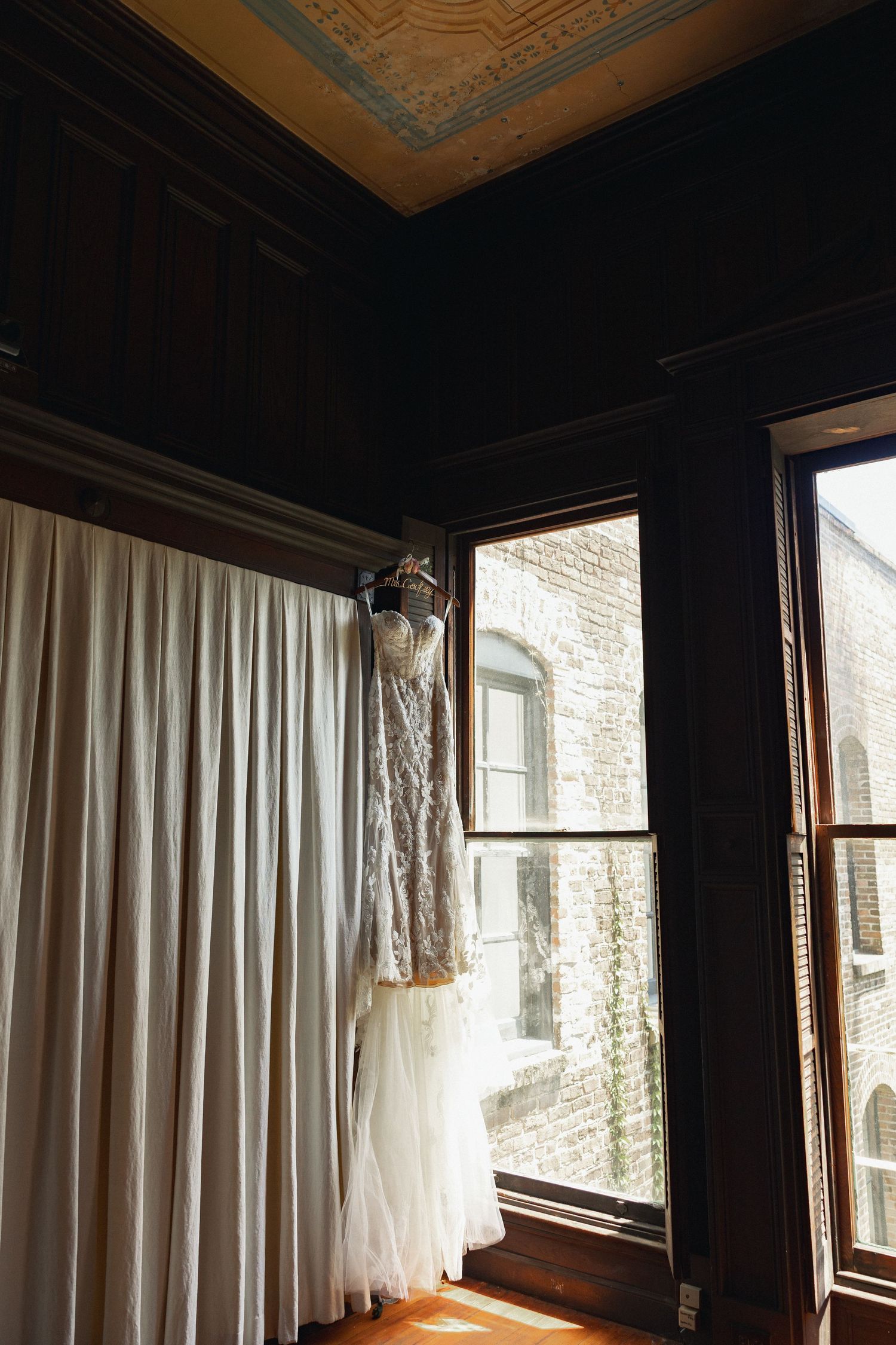 Elegant lace wedding dress hanging near a window in a dark wood-paneled room with ornate ceiling details.