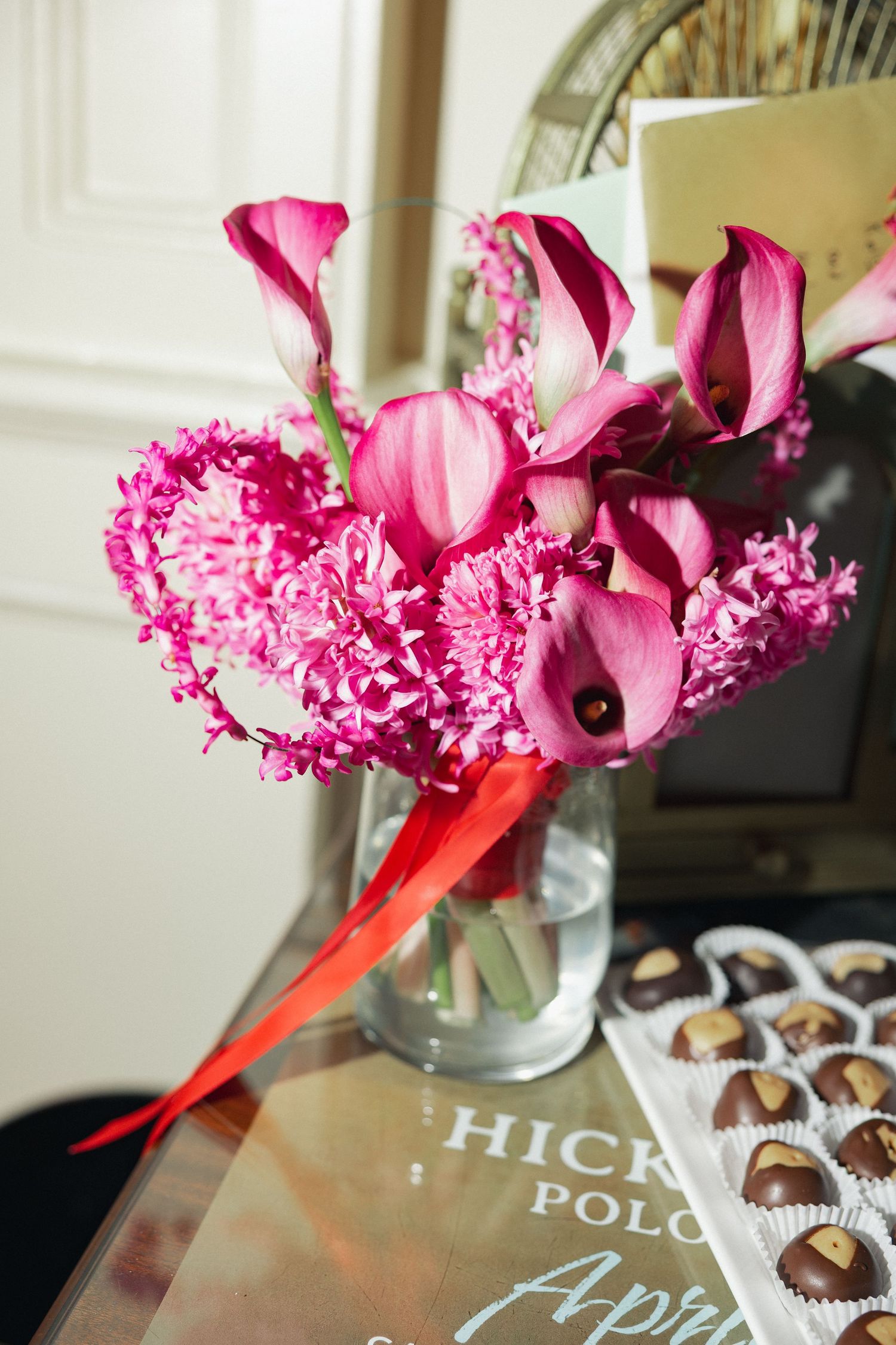 Pink calla lilies and hyacinths in glass vase with red ribbon beside Hickey polo book and chocolate box.