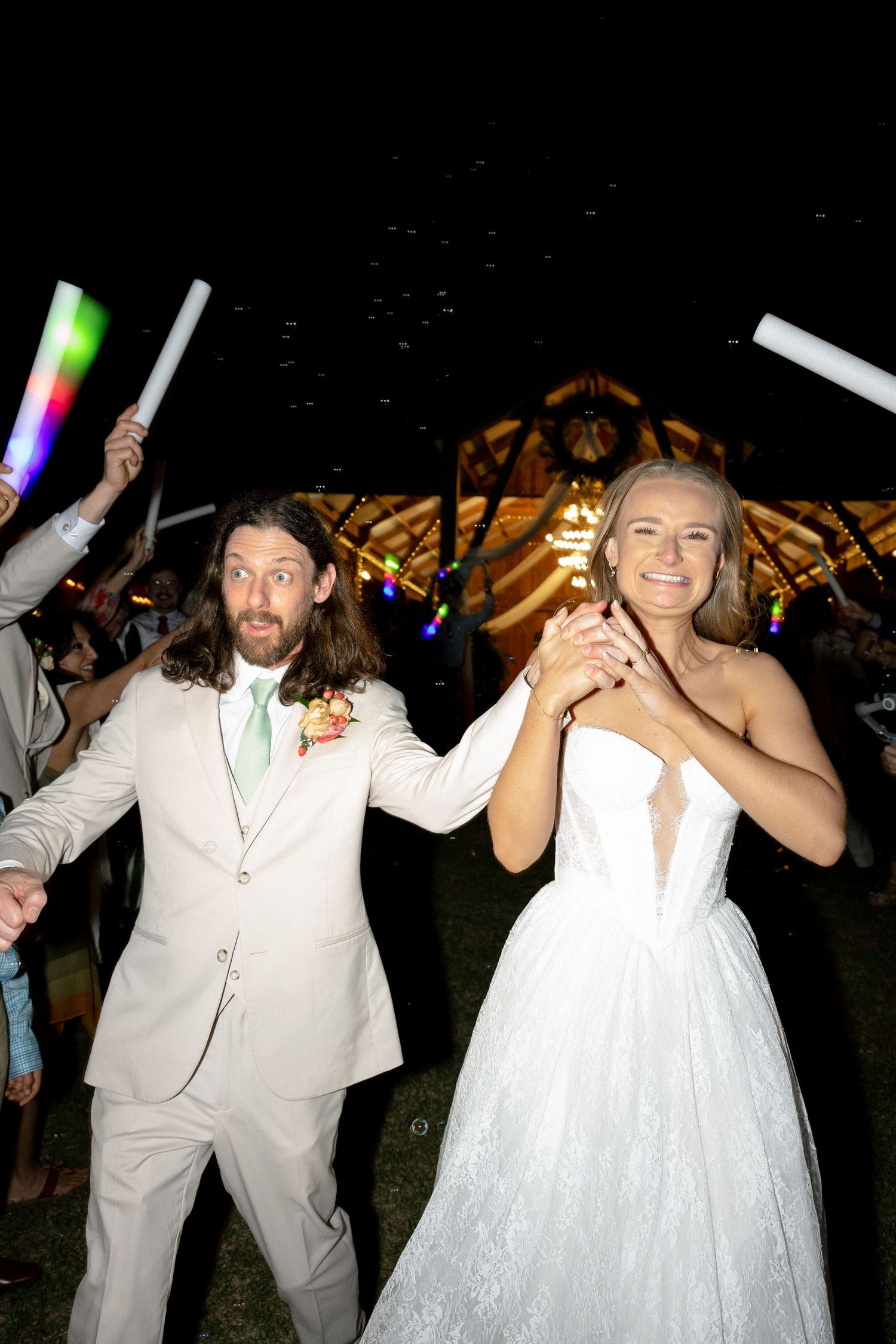 Bride in white gown and groom in tan suit celebrate wedding exit with colorful glow sticks at night.