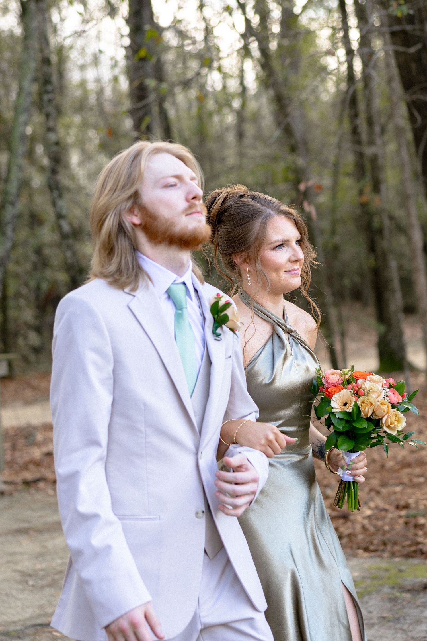 A long-haired groomsman in white suit and mint tie escorts a bridesmaid in olive satin gown at outdoor wedding ceremony.