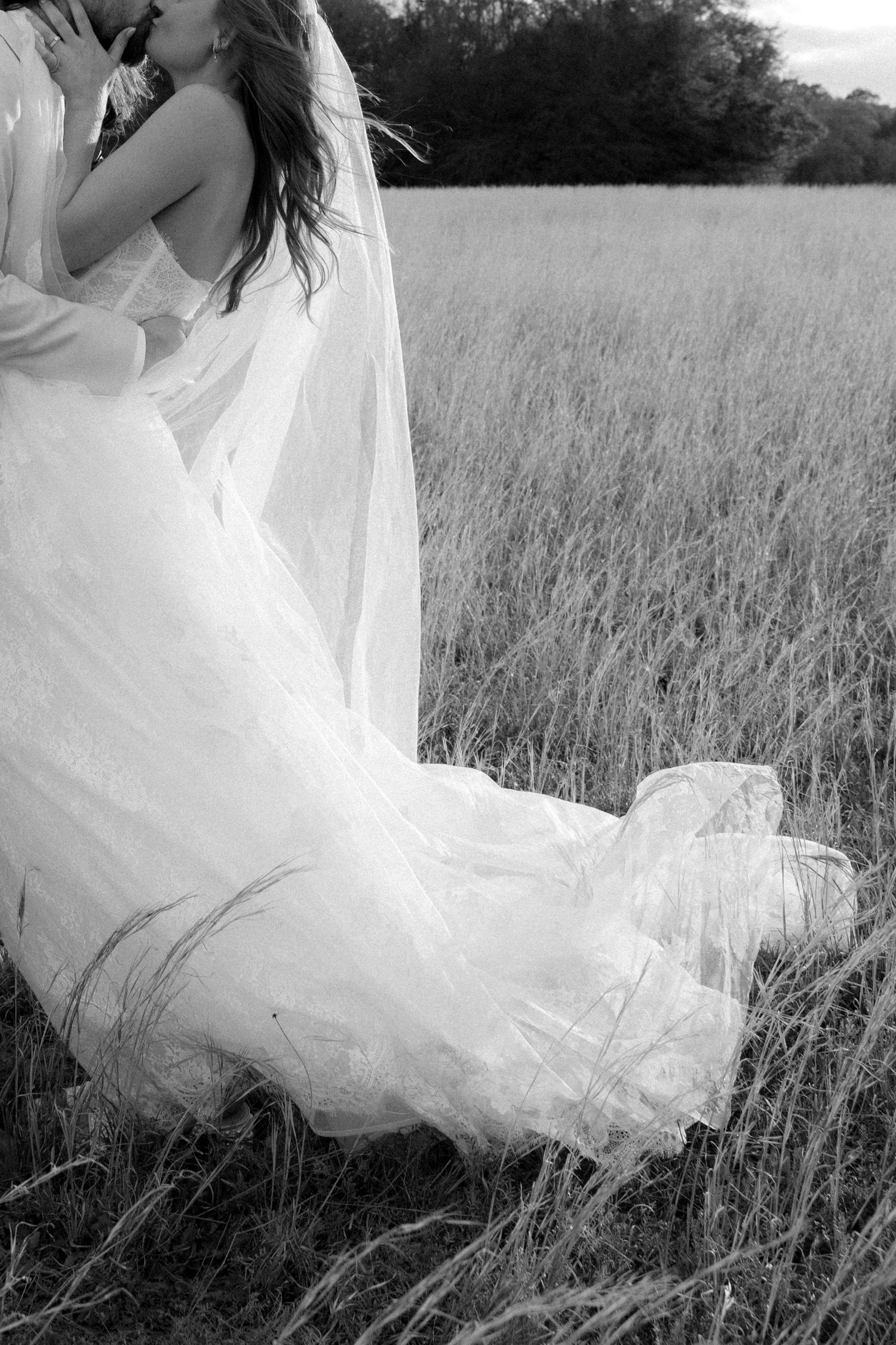 Black and white artistic shot of bride in flowing lace gown and veil in open field.