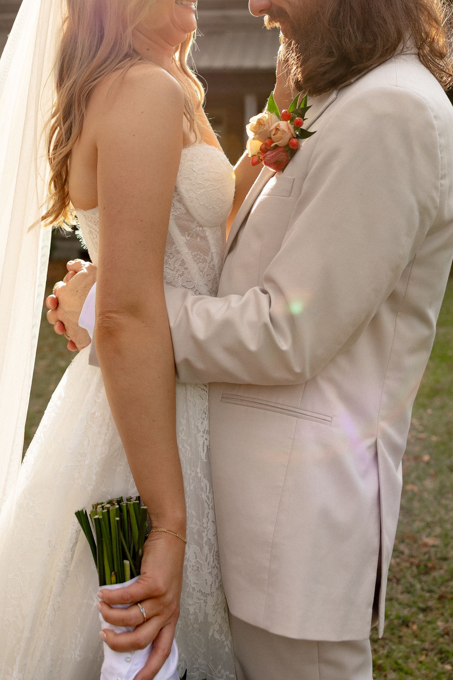 Close-up of bride and groom standing together, groom in beige suit with floral boutonniere, bride holding bouquet.