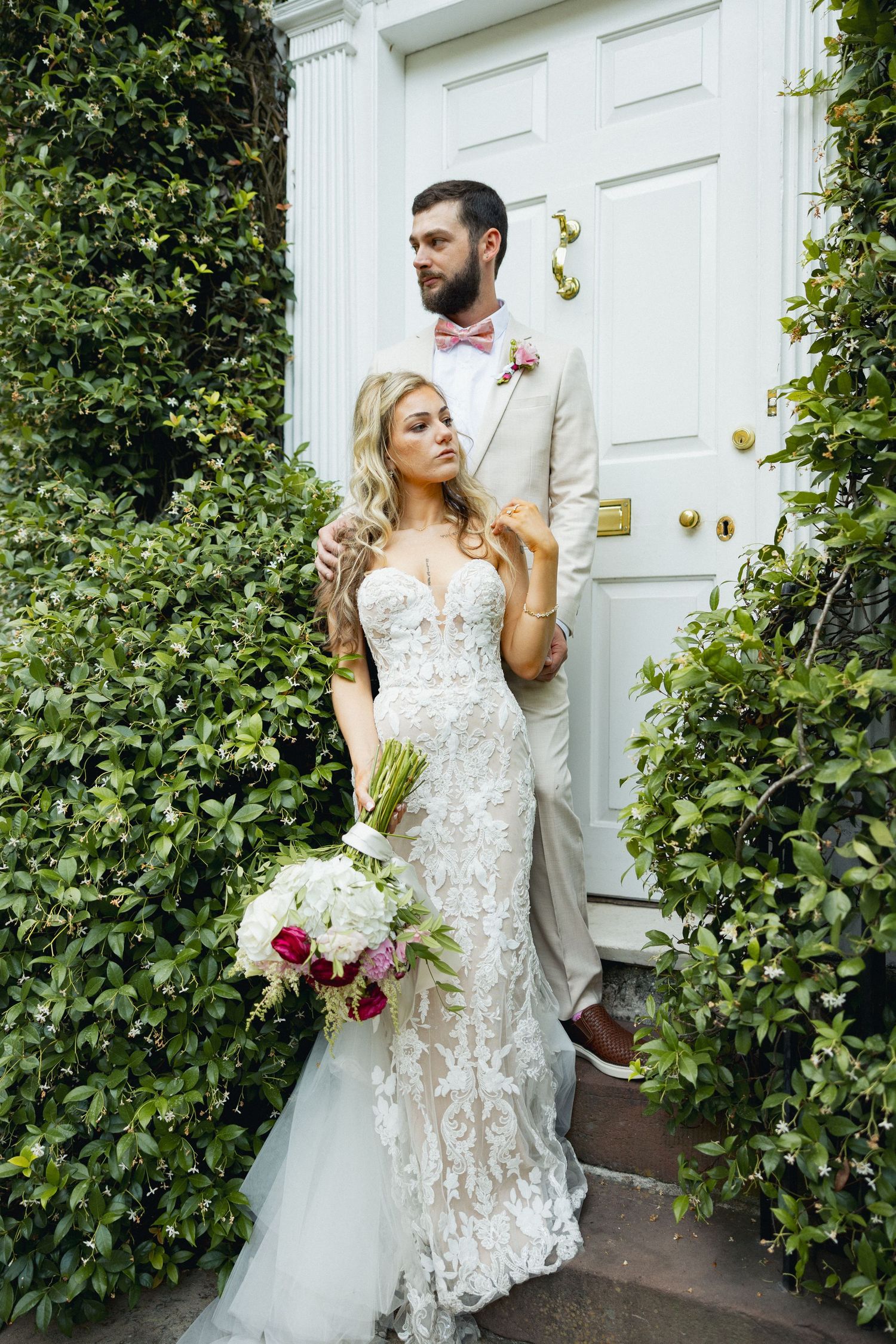 Bride in lace gown holding bouquet stands with bearded groom in tan suit before white door surrounded by greenery.