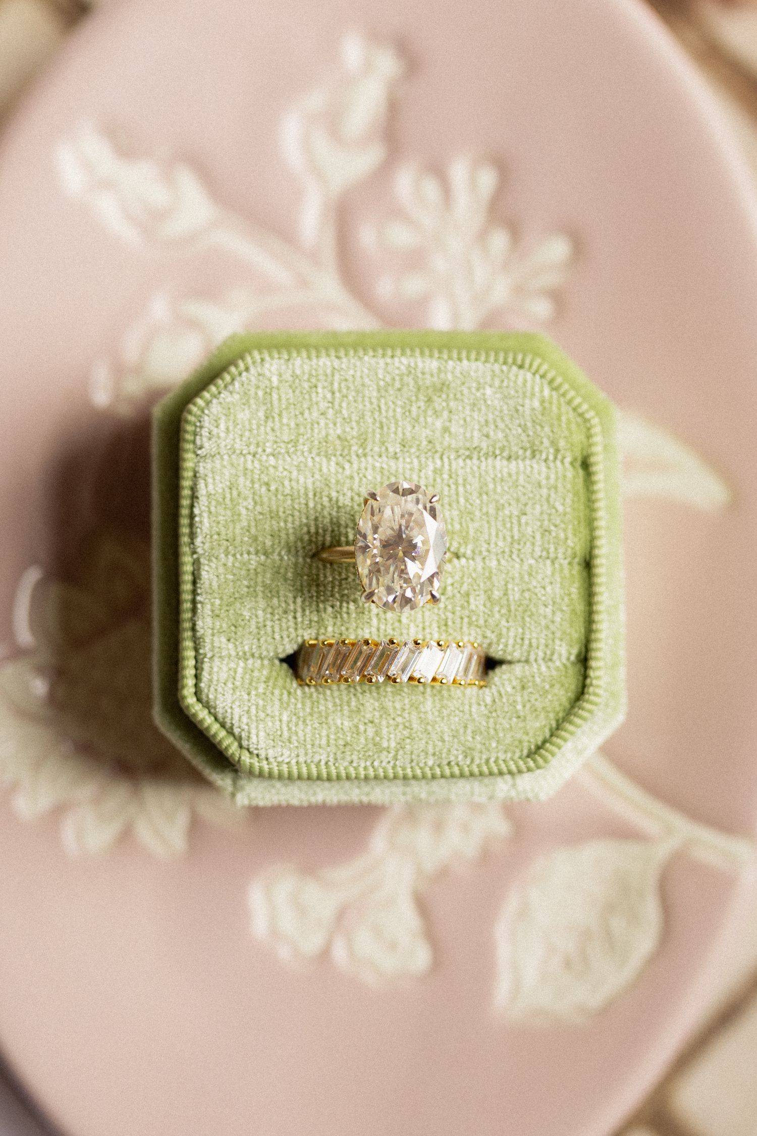Oval diamond engagement ring with gold baguette wedding band displayed in a green velvet ring box on a pink floral plate.