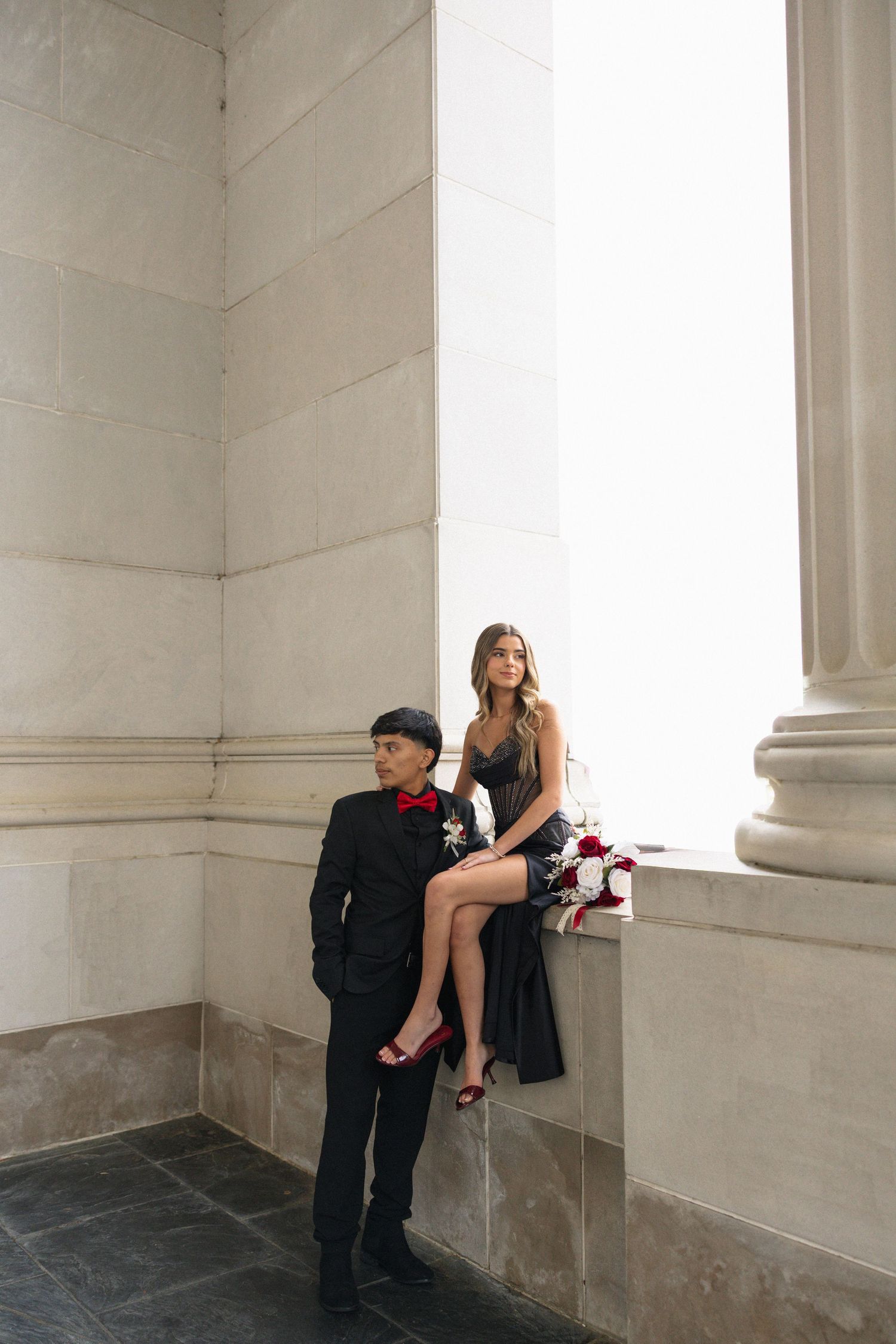 Elegant couple in black formal wear posing at marble columns for prom photos.