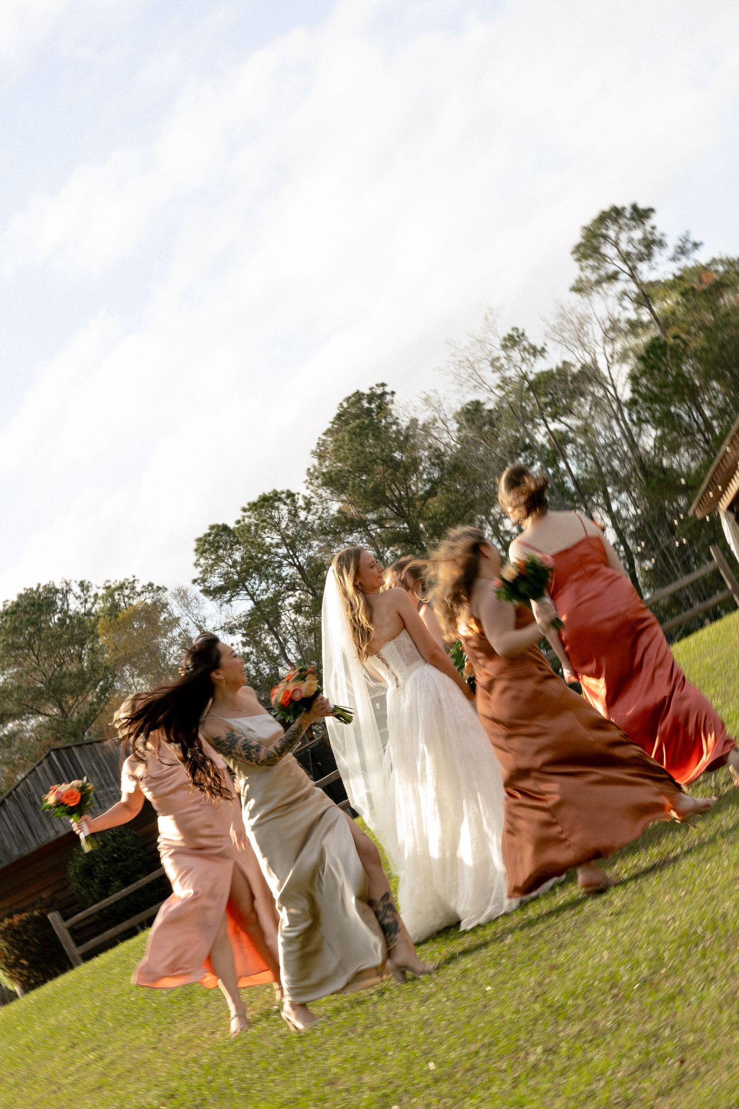 Bride and bridesmaids in colorful dresses running and dancing joyfully on green lawn at outdoor wedding.