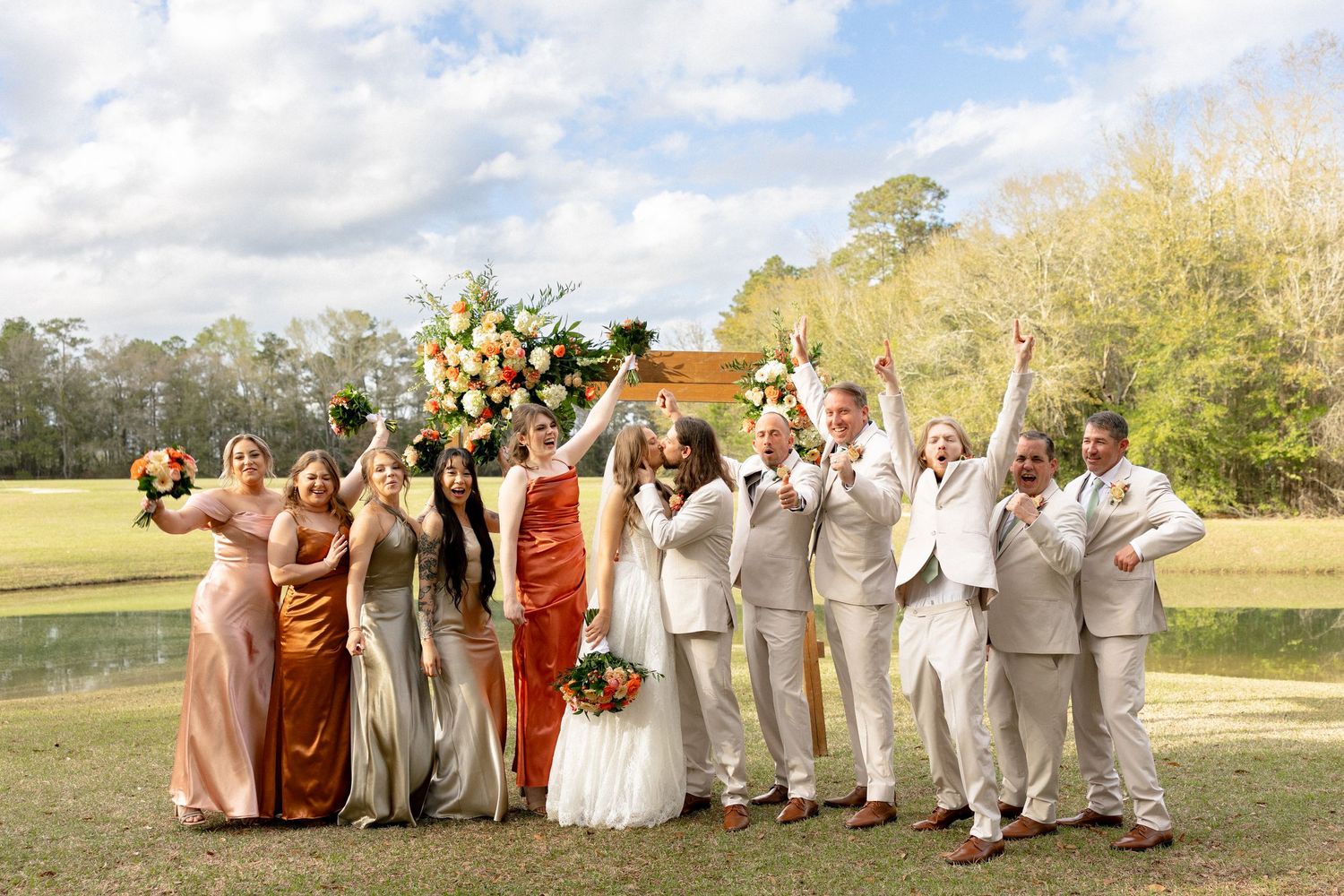 Excited wedding party celebrates with raised arms under floral arch on green lawn.