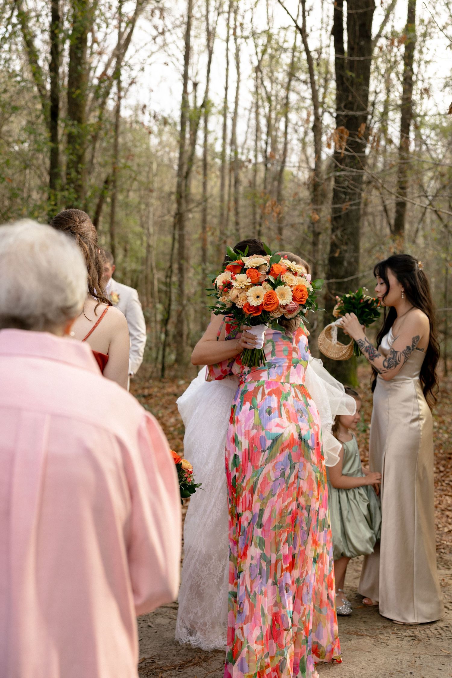 Wedding guests gather around bride in wooded outdoor setting, woman in colorful dress holding orange bouquet.