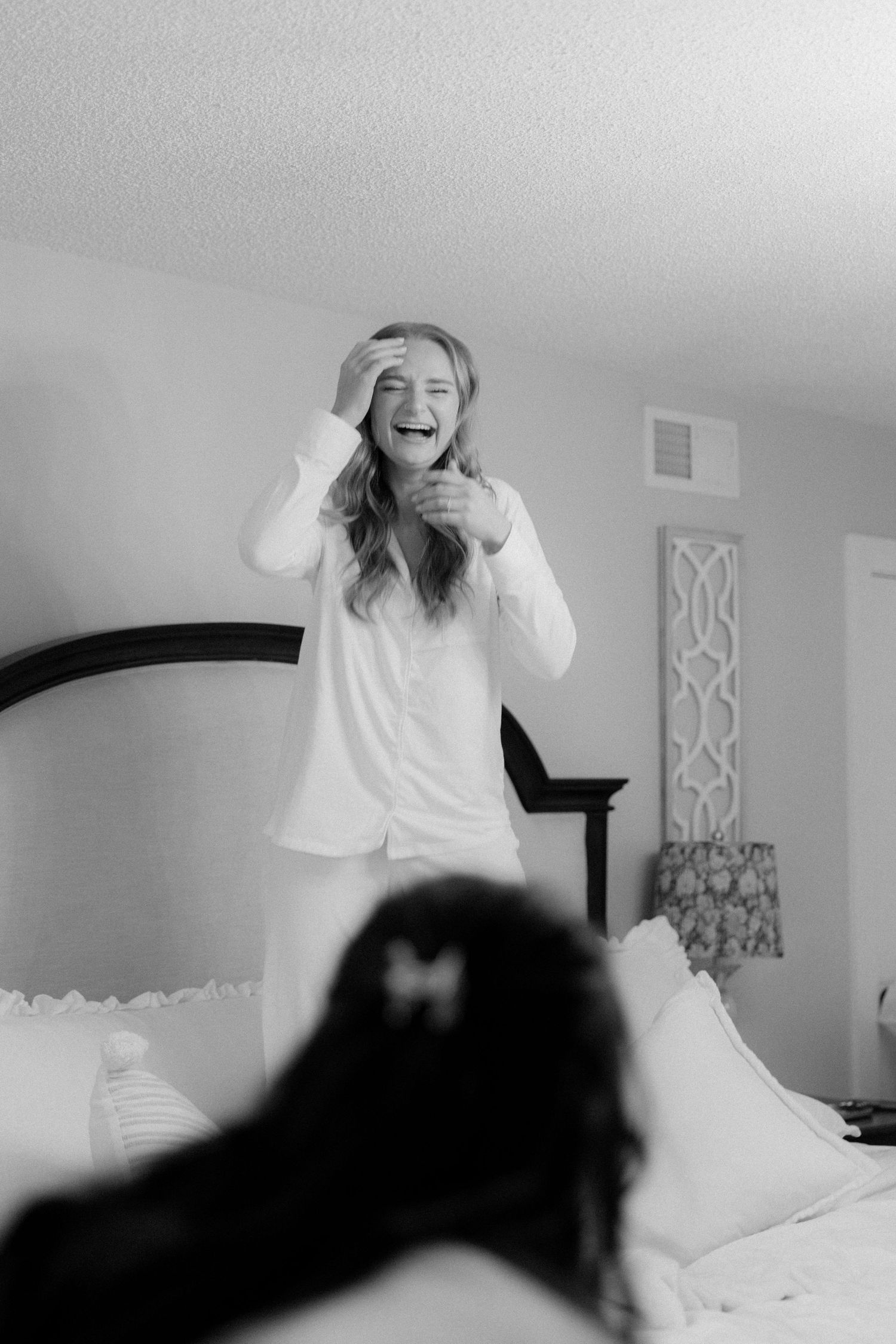 Laughing bride in white pajamas standing on bed during wedding morning preparations.