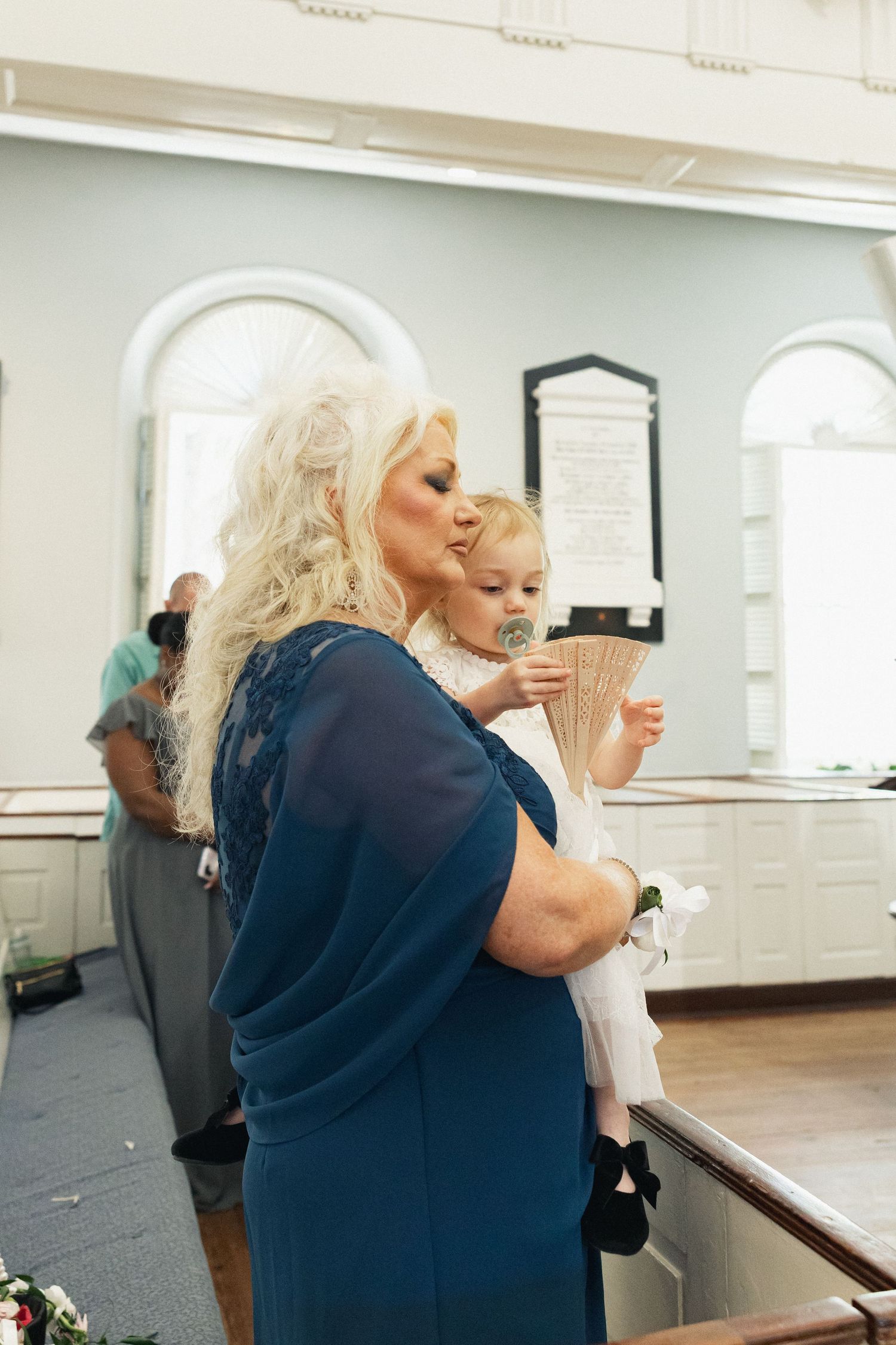 A blonde woman in a blue dress holds a young flower girl with a fan during a wedding ceremony.