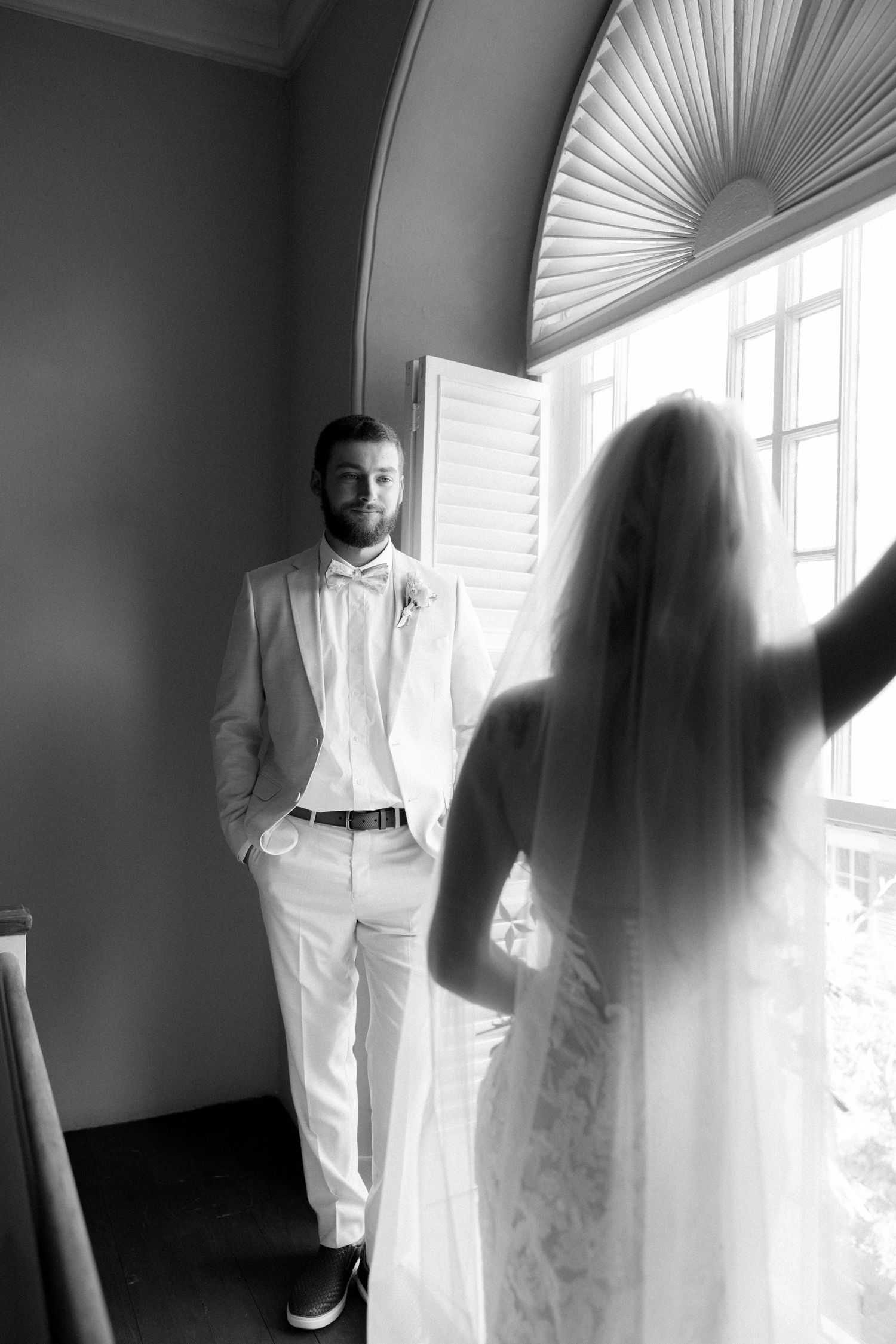 Groom in white suit smiles at bride in wedding dress standing by arched window in black and white photo.