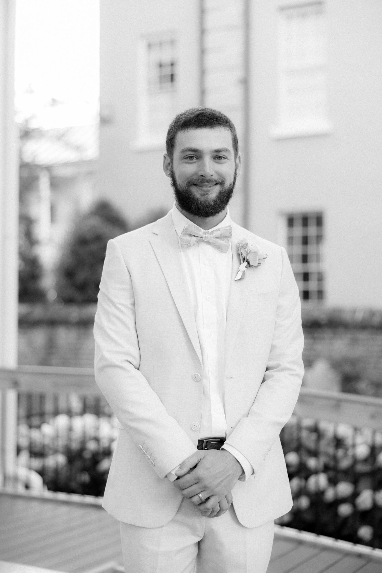 Smiling groom in white suit with bow tie and boutonniere posing outdoors in black and white photo.