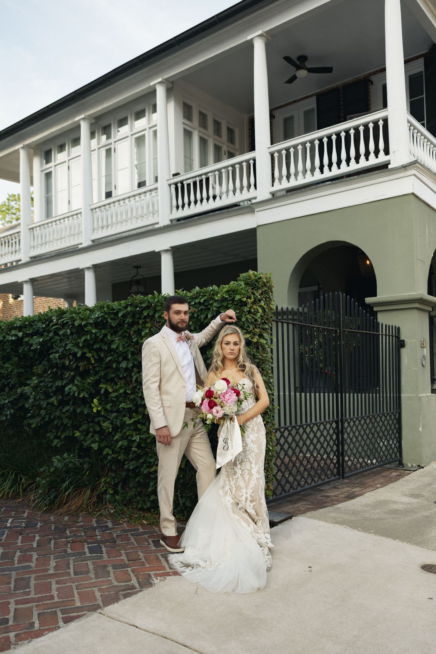 Elegant bride and groom stand before grand white Southern plantation-style home with double porches and iron gate.