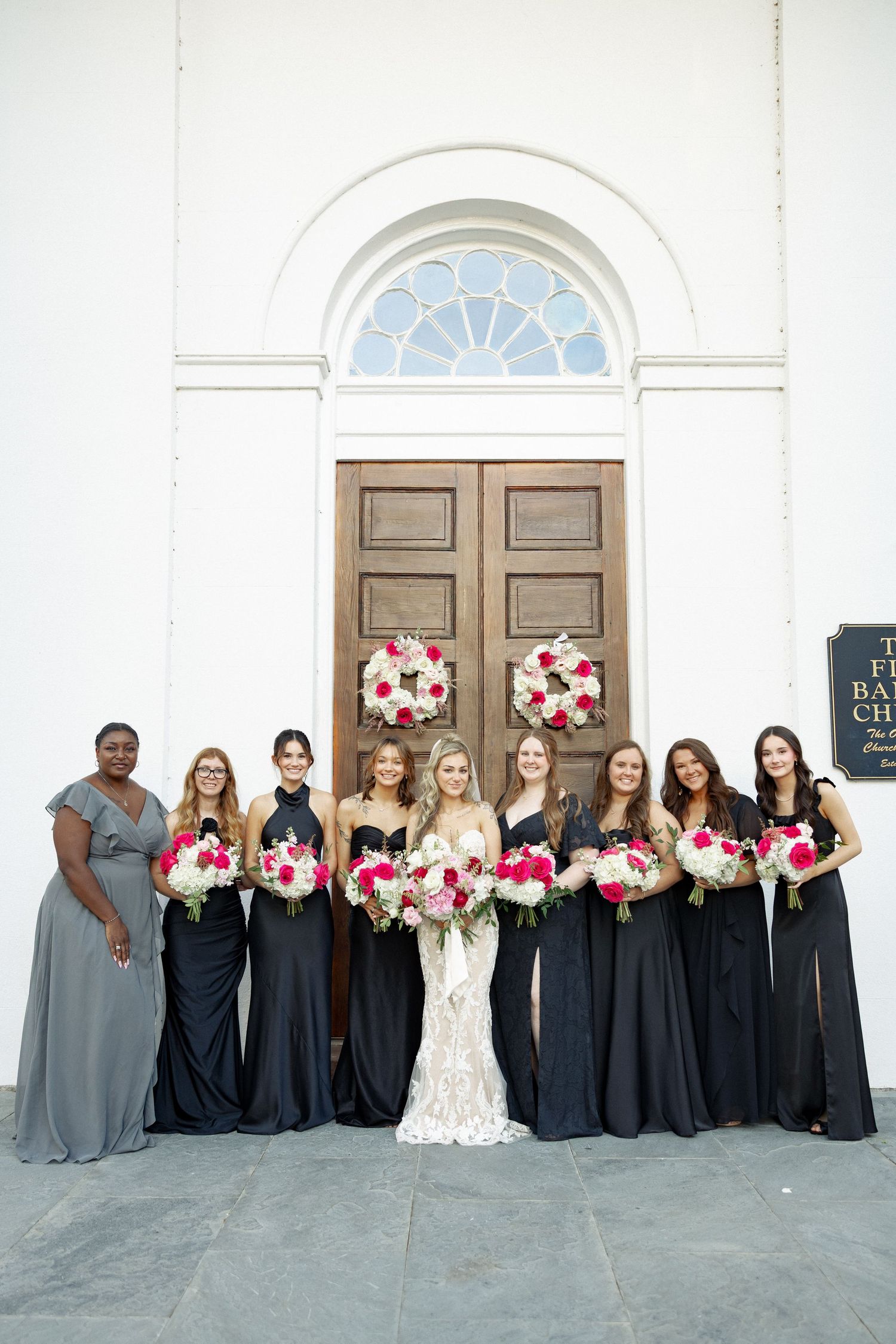 Bride in lace gown with bridesmaids in navy dresses holding pink and white bouquets outside First Baptist Church.