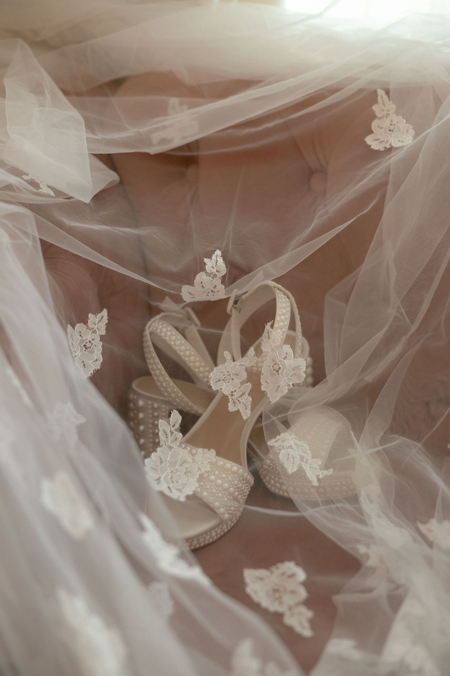 Elegant bridal wedding shoes with lace floral details and pearl accents resting on a delicate white tulle veil.