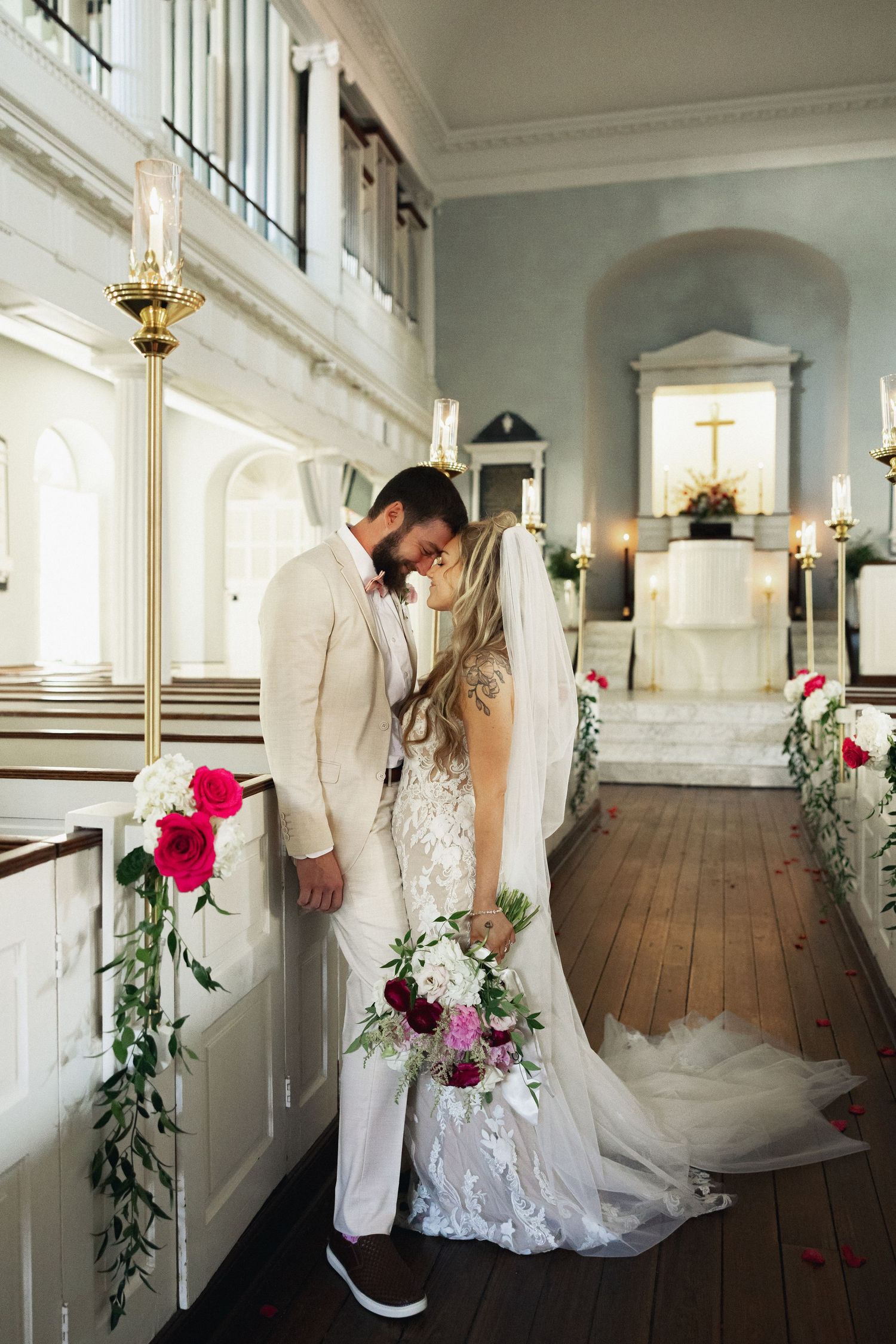 Bride and groom share a romantic kiss in a elegant white church adorned with red and white floral arrangements.