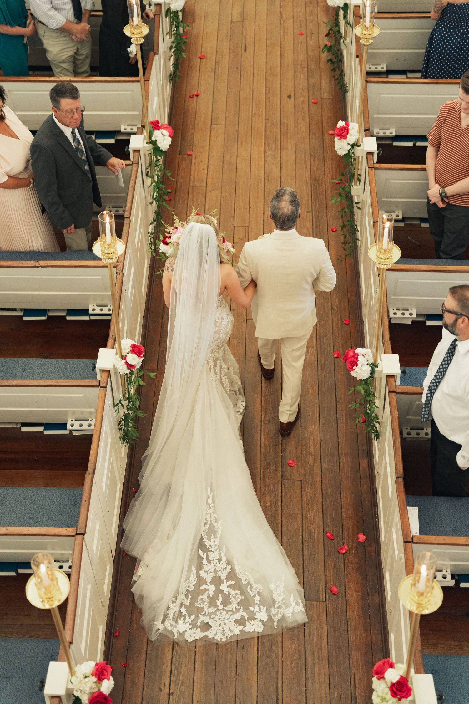 Aerial view of bride in lace gown and father walking down church aisle adorned with floral garlands.