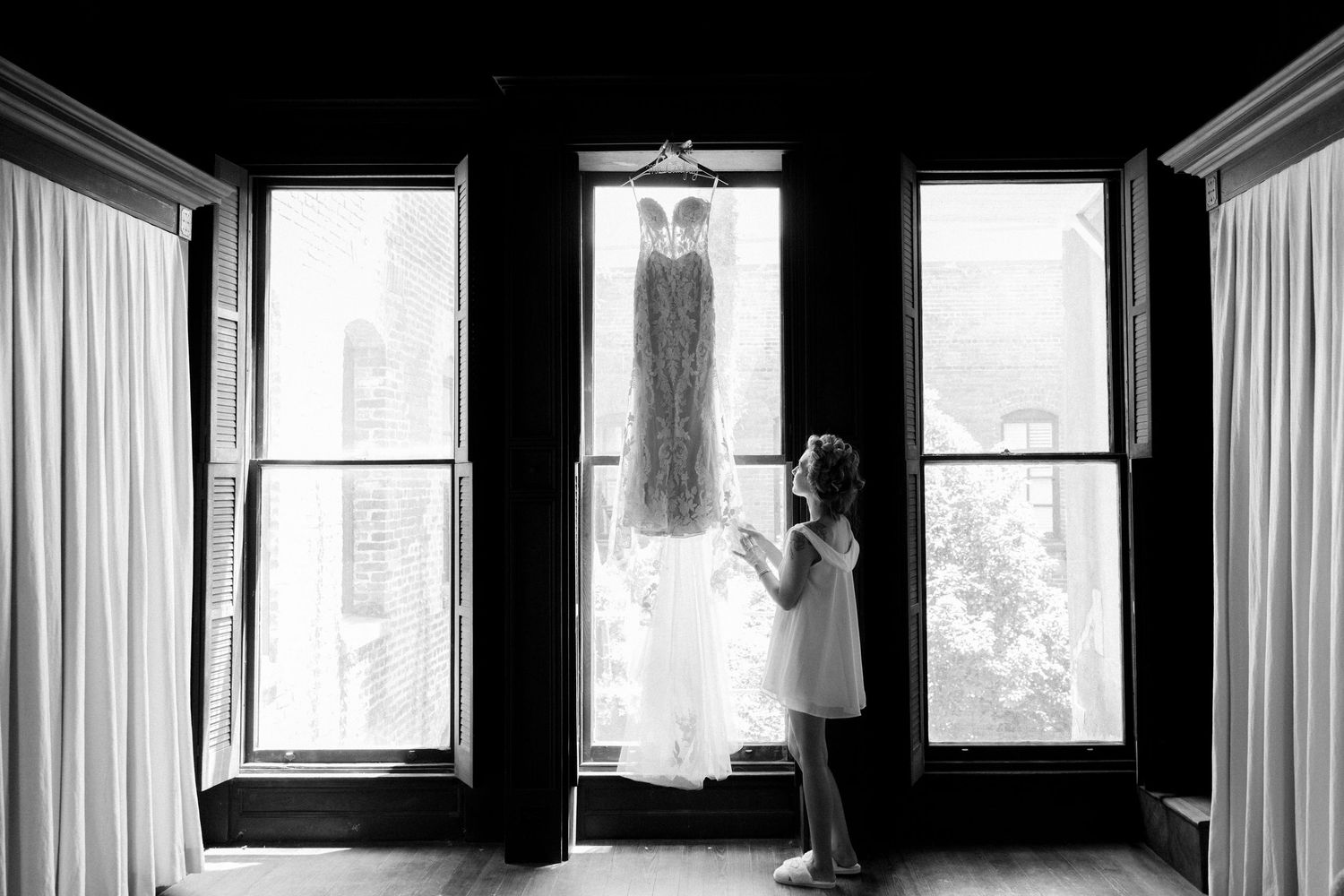 Black and white photo of a flower girl admiring a wedding dress hanging in a sunlit window.