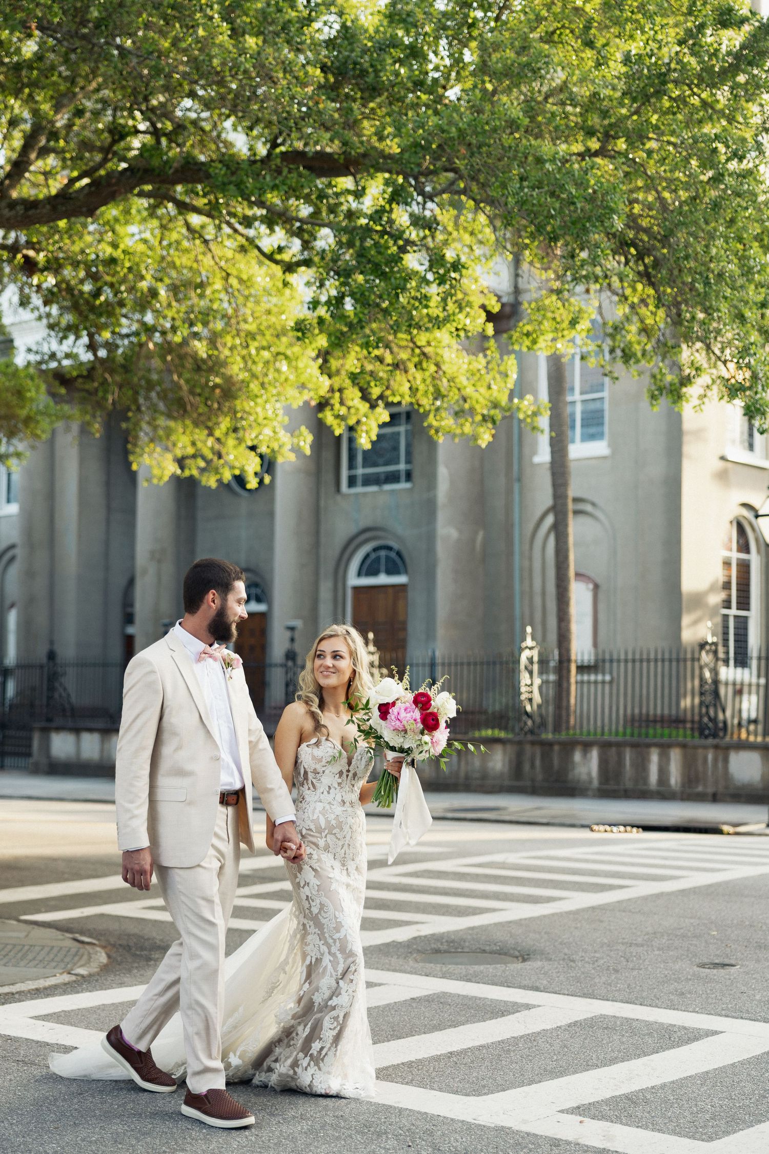 Bride in lace gown and groom in beige suit walk hand-in-hand across a crosswalk near a historic church.