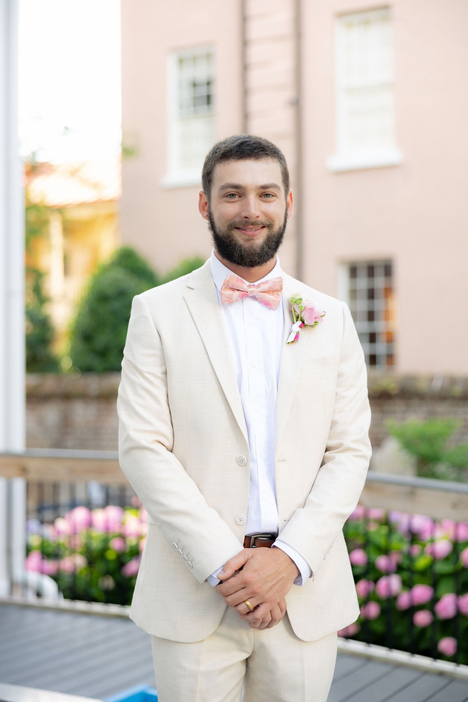 Bearded groom smiles in cream tuxedo with pink bow tie and boutonniere on outdoor terrace.